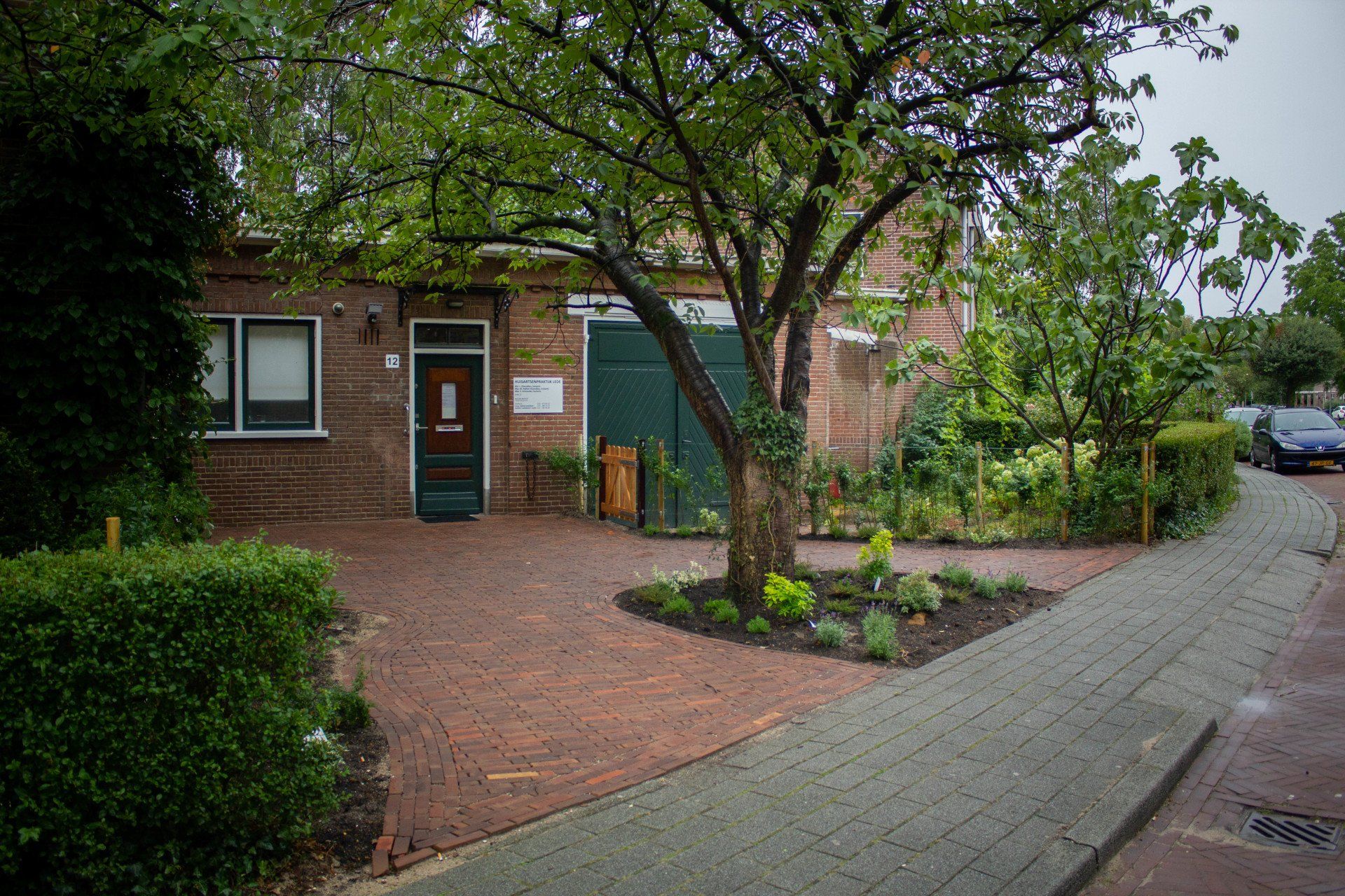 Bakstenen gebouw met groene deur en garage, boom ervoor, bakstenen en grijs bestraat terrein, bewolkte dag.