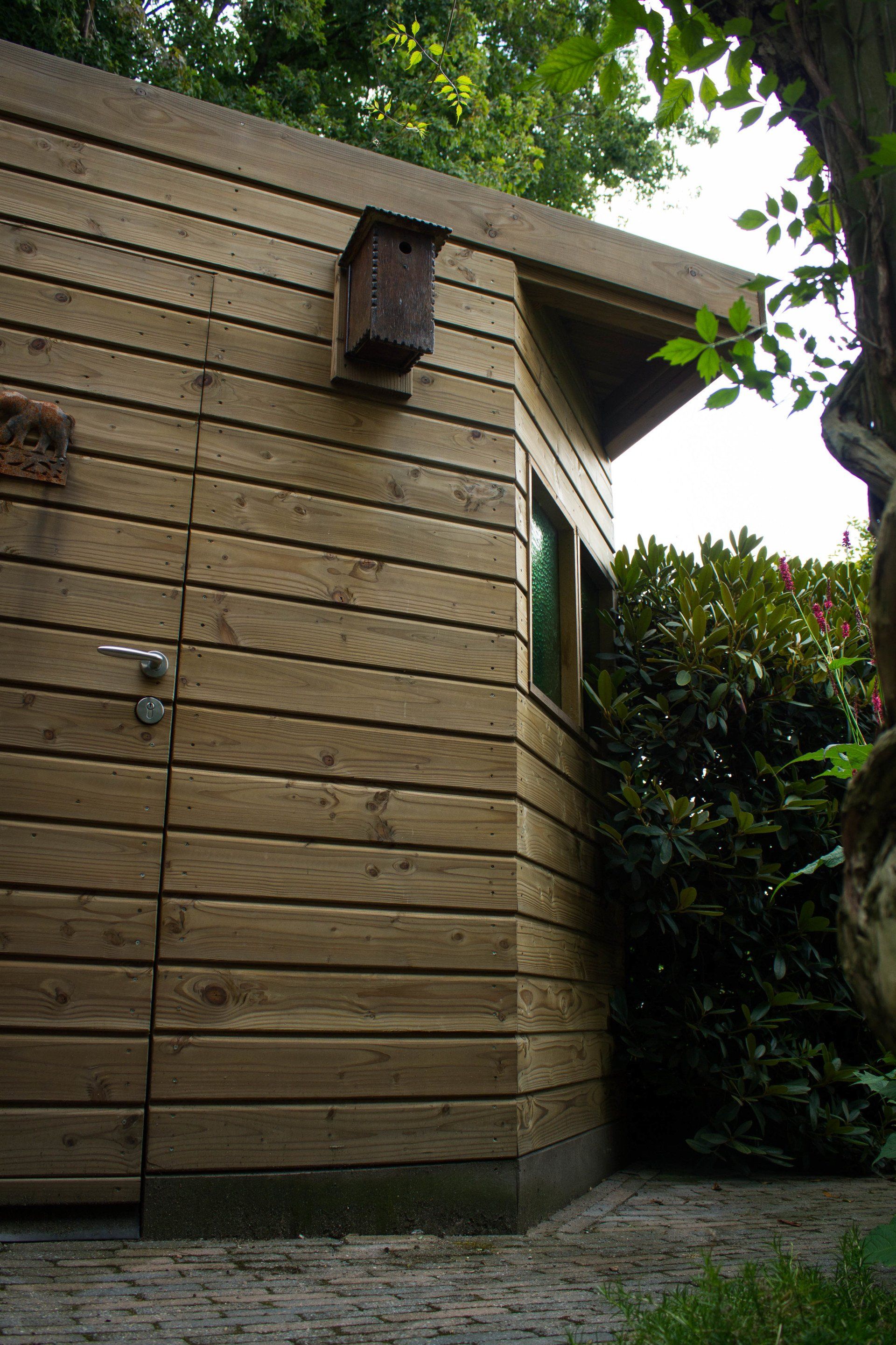 Houten schuur met vogelhuisje, gelegen in een tuin met groen en een stenen pad.