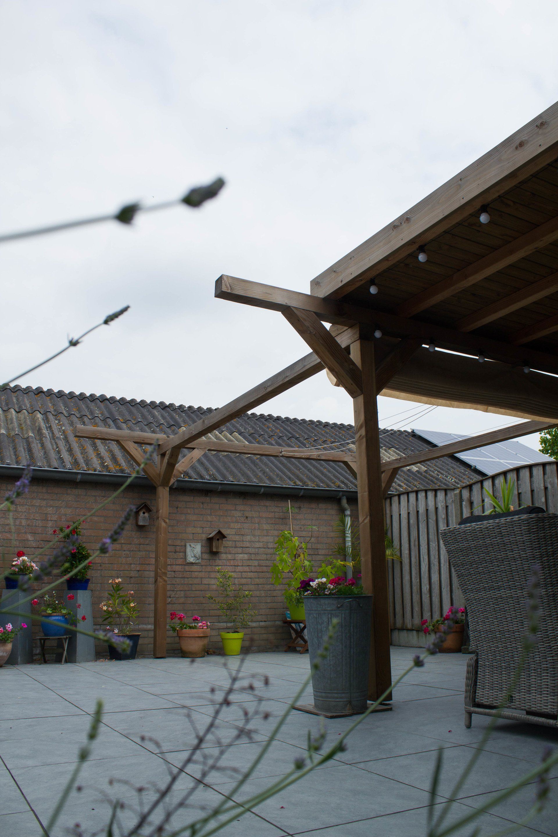 Houten pergola boven een terras, bakstenen gebouw op de achtergrond, bewolkte lucht.