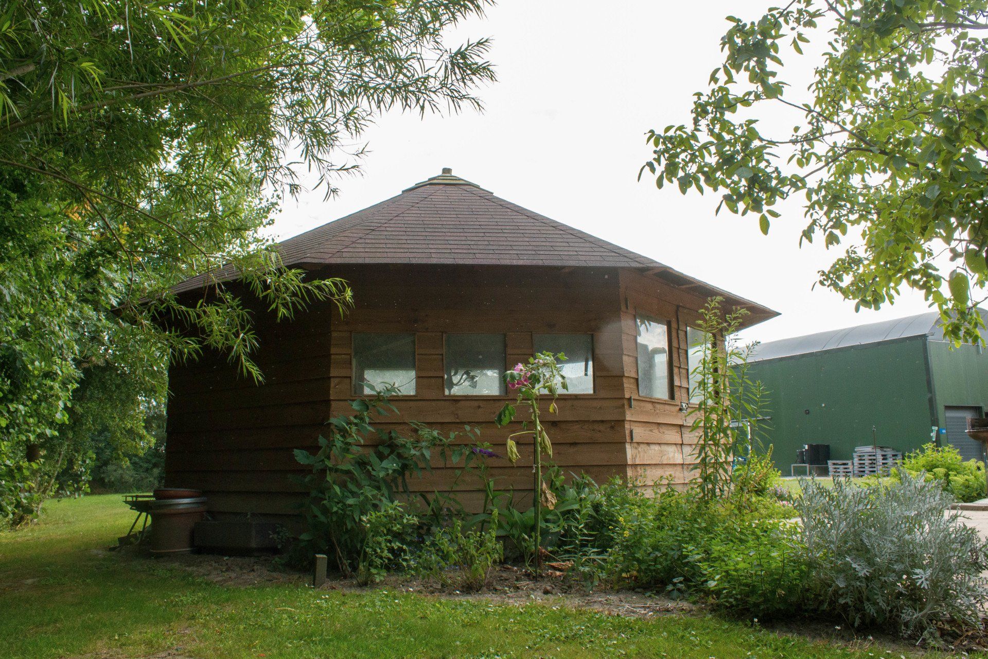 Houten zeshoekig tuinpaviljoen met ramen, op een grasveld, omgeven door bomen.