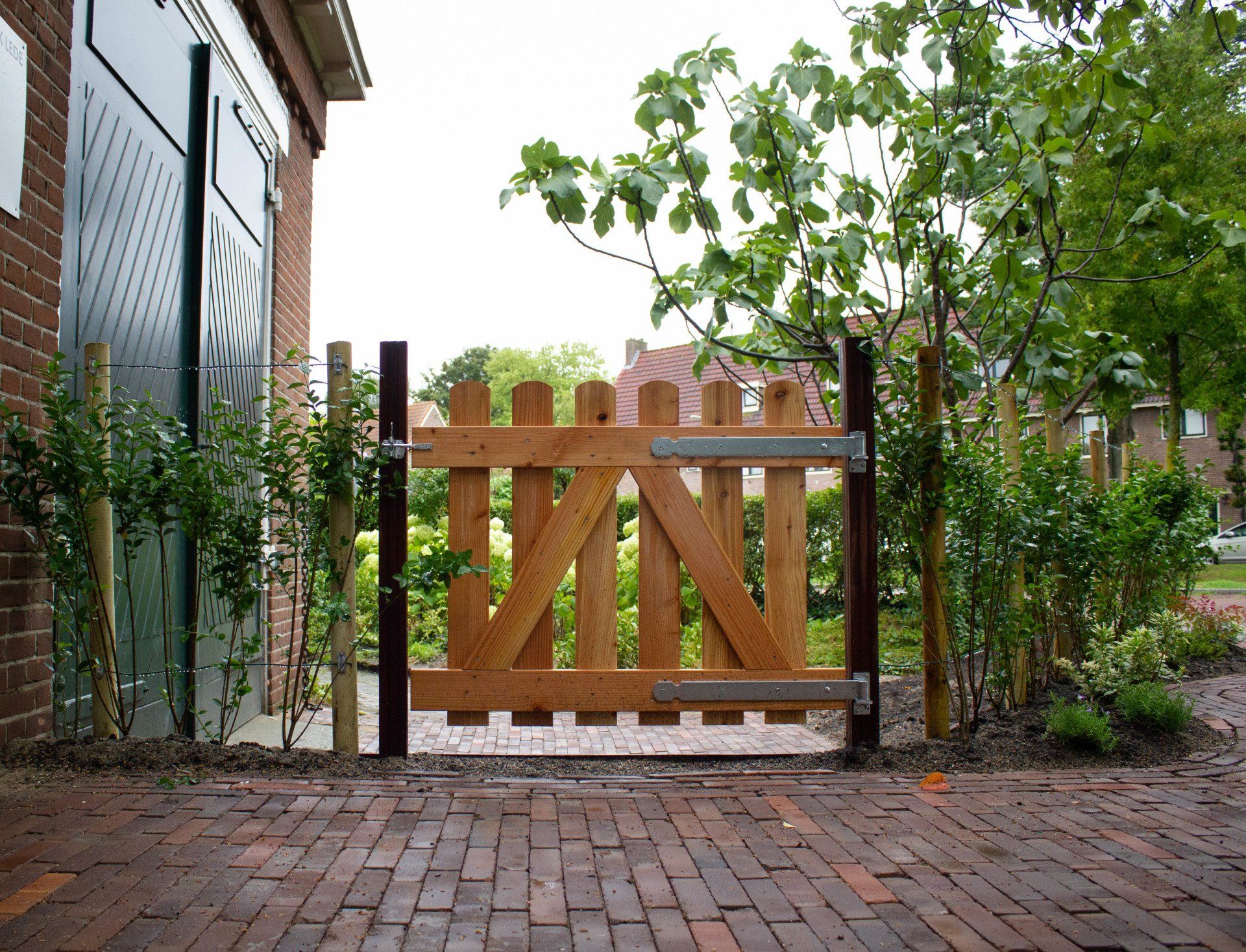 Houten poort met een diagonale versteviging, geplaatst in een bakstenen pad, tussen metalen palen.