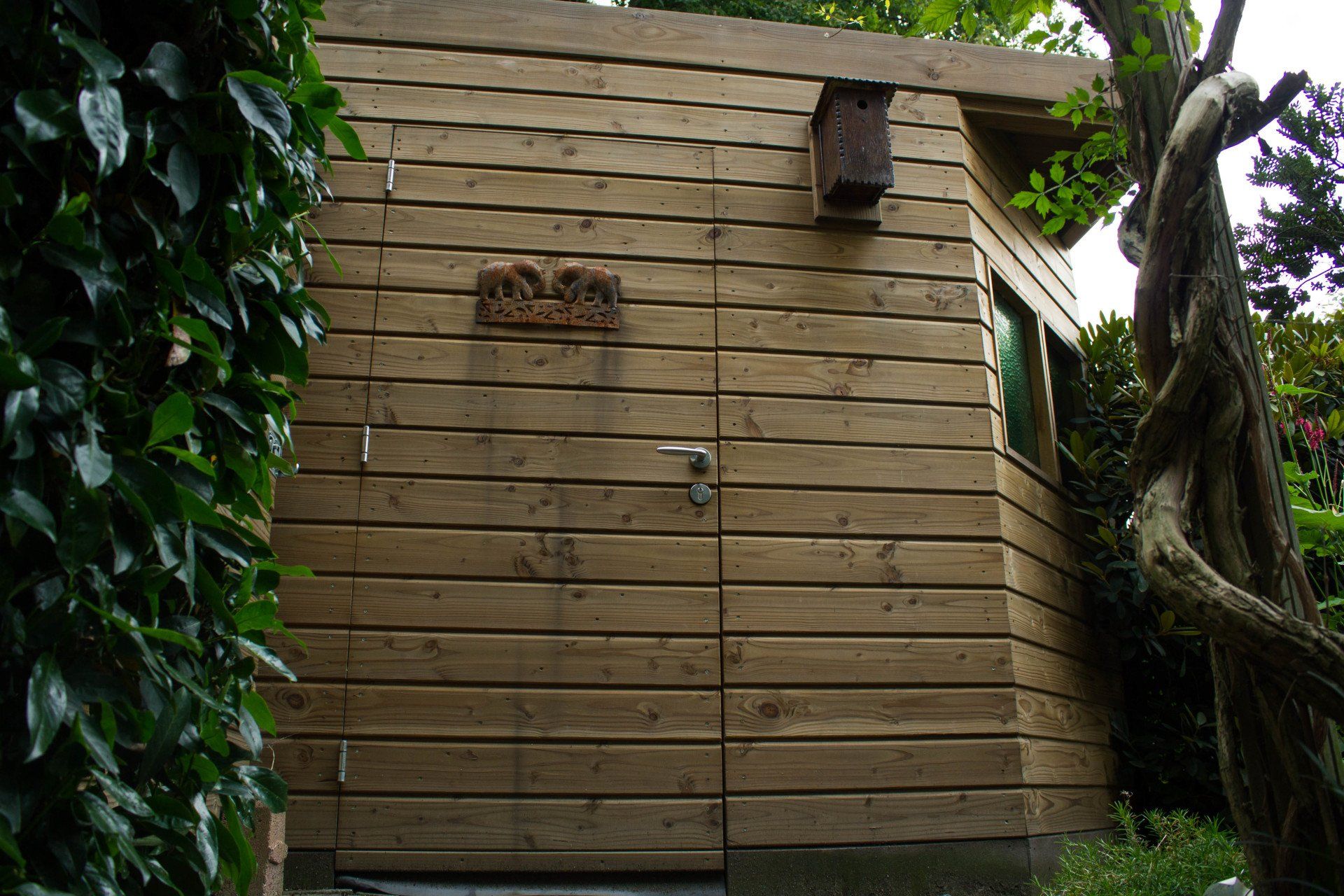 Houten schuur met een vogelhuisje, een plaquette en een boom met bladeren aan de rechterkant. Groene bladeren aan de linkerkant.