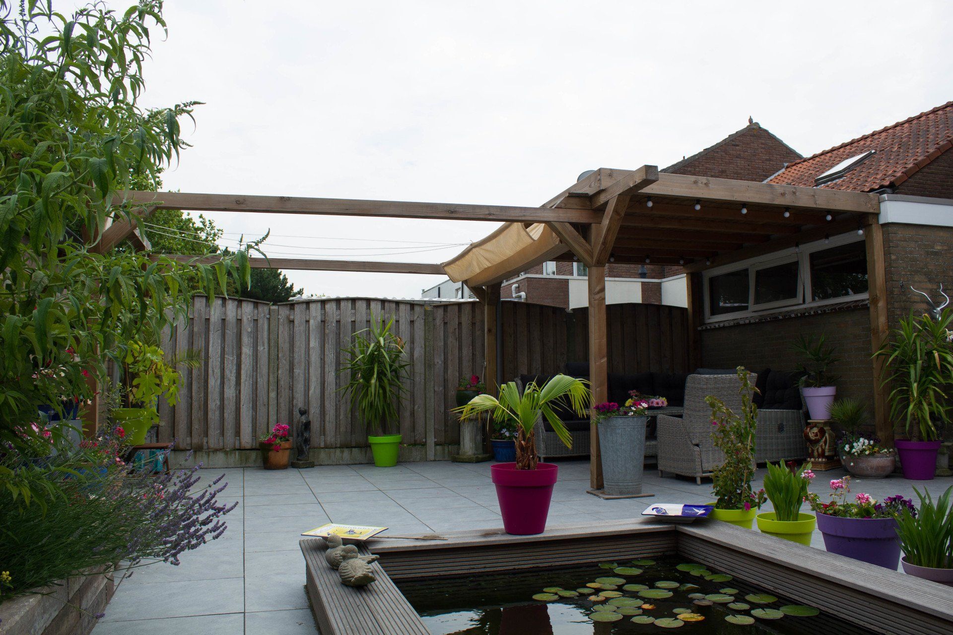 Achtertuin met een houten pergola, terras, vijver en potplanten.