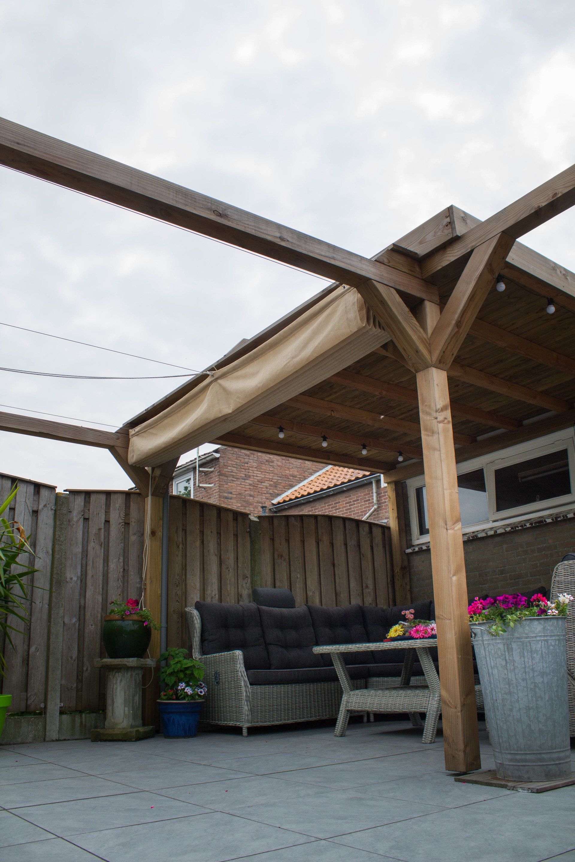 Houten pergola boven een zithoek buiten; met een bank, tafel en potplanten.