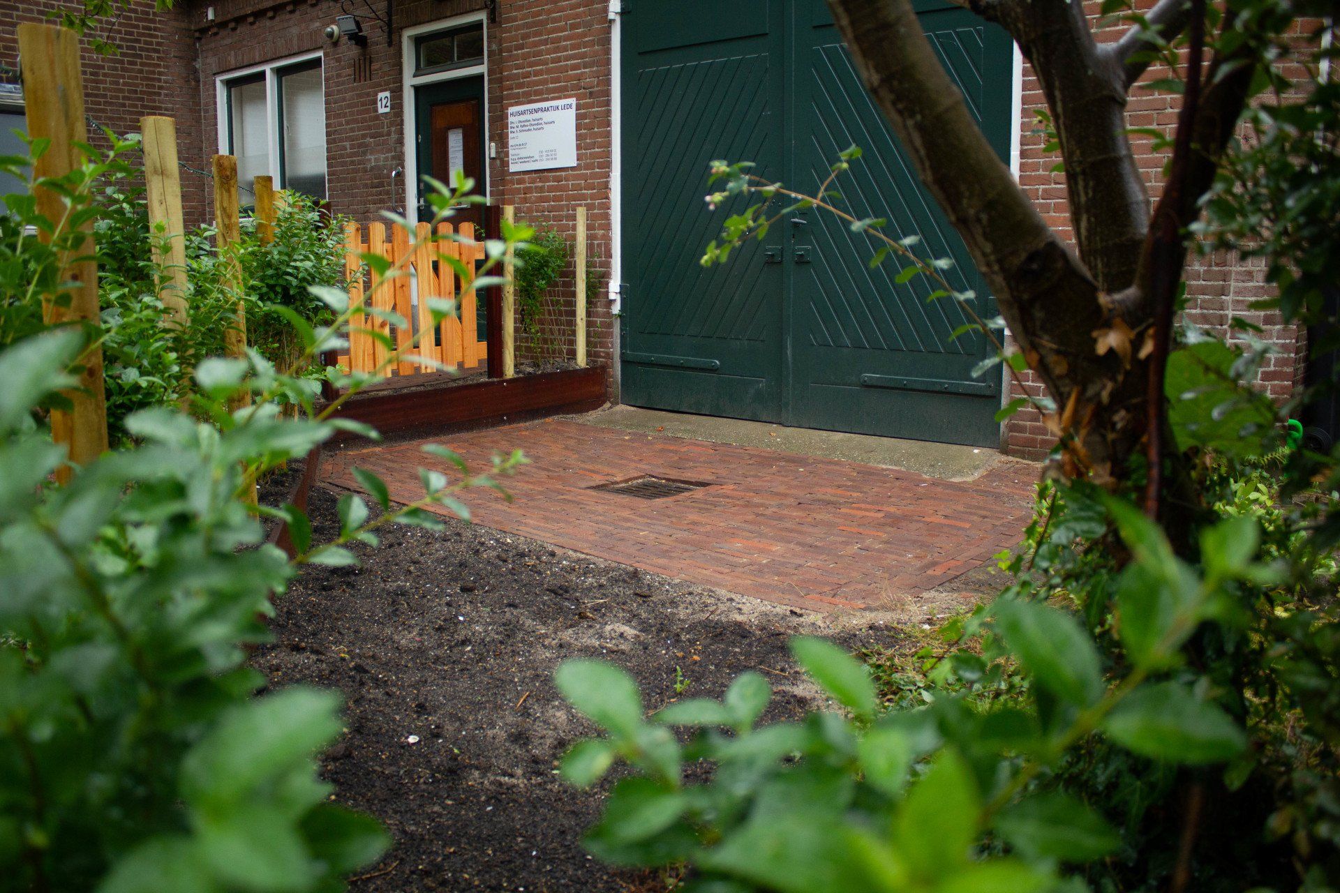 Ingang met rode bakstenen bestrating en groene garagedeur, omgeven door een tuin met een houten schutting.