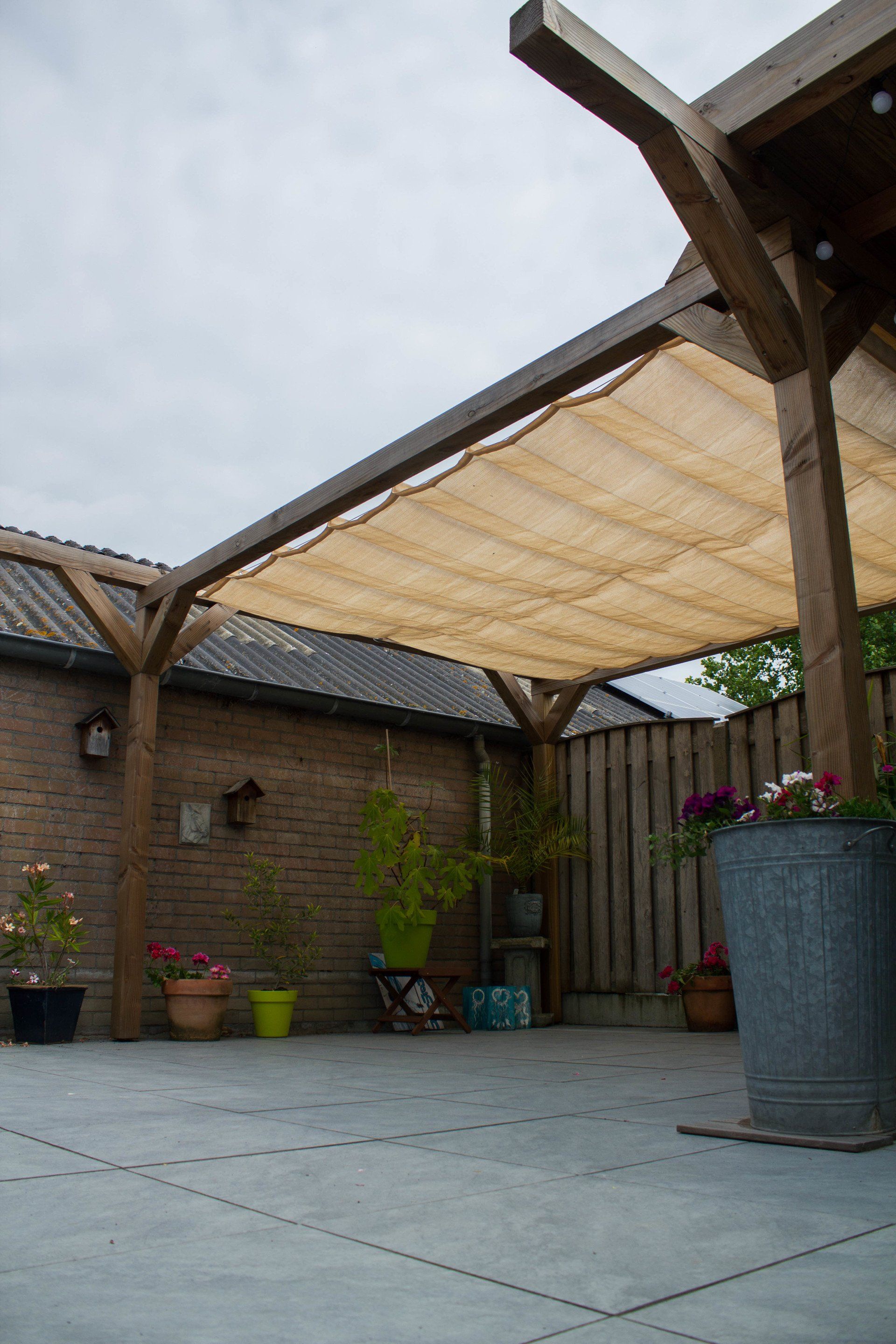Een houten pergola boven een terras, met schaduwdoek en potplanten langs een muur en schutting.