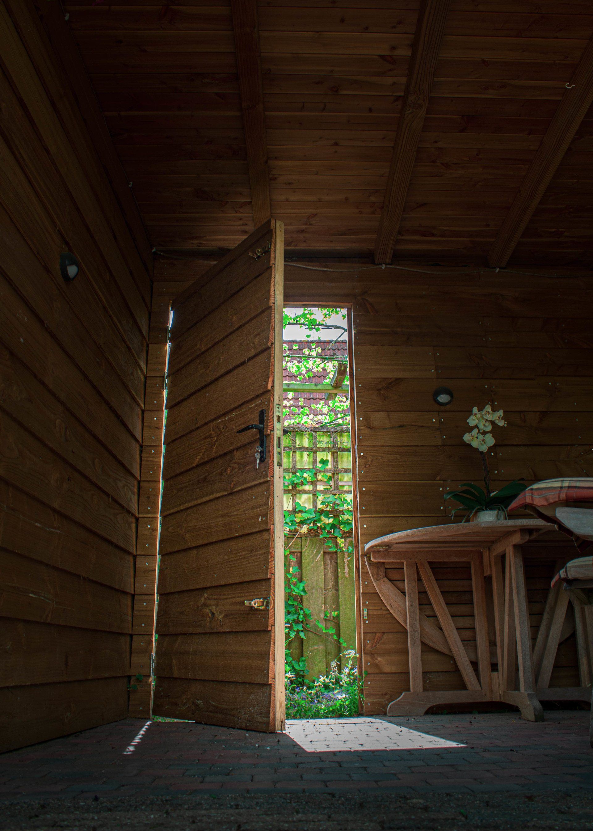 Interieur van een houten schuur, deur op een kier, uitzicht op de zonnige tuin, tafel en krukjes binnen.