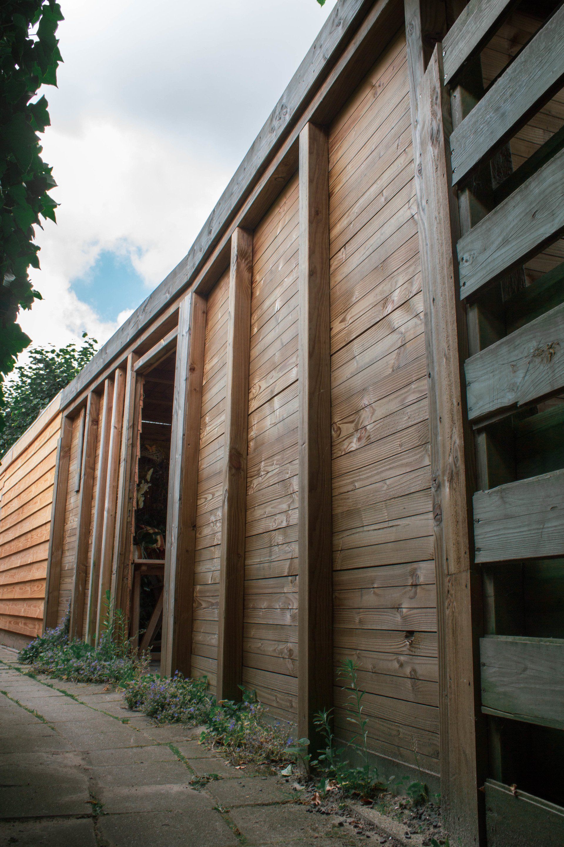 Houten constructie met verticale balken en planken, mogelijk een schuur of gebouw, met begroeiing aan de voet.