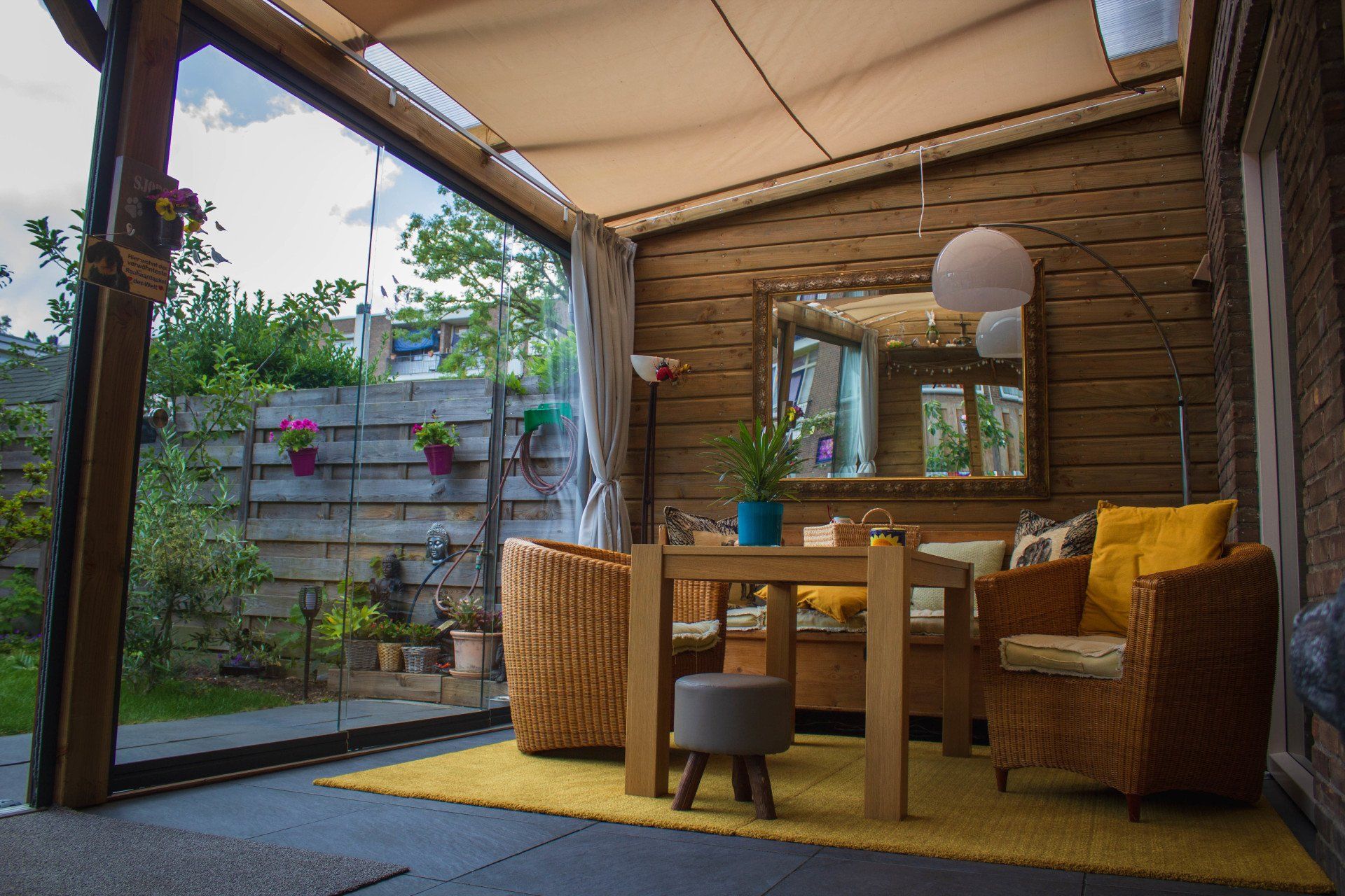 Overdekt terras met houten wanden en meubilair, geel vloerkleed, tafel, rieten stoelen en uitzicht op de tuin.