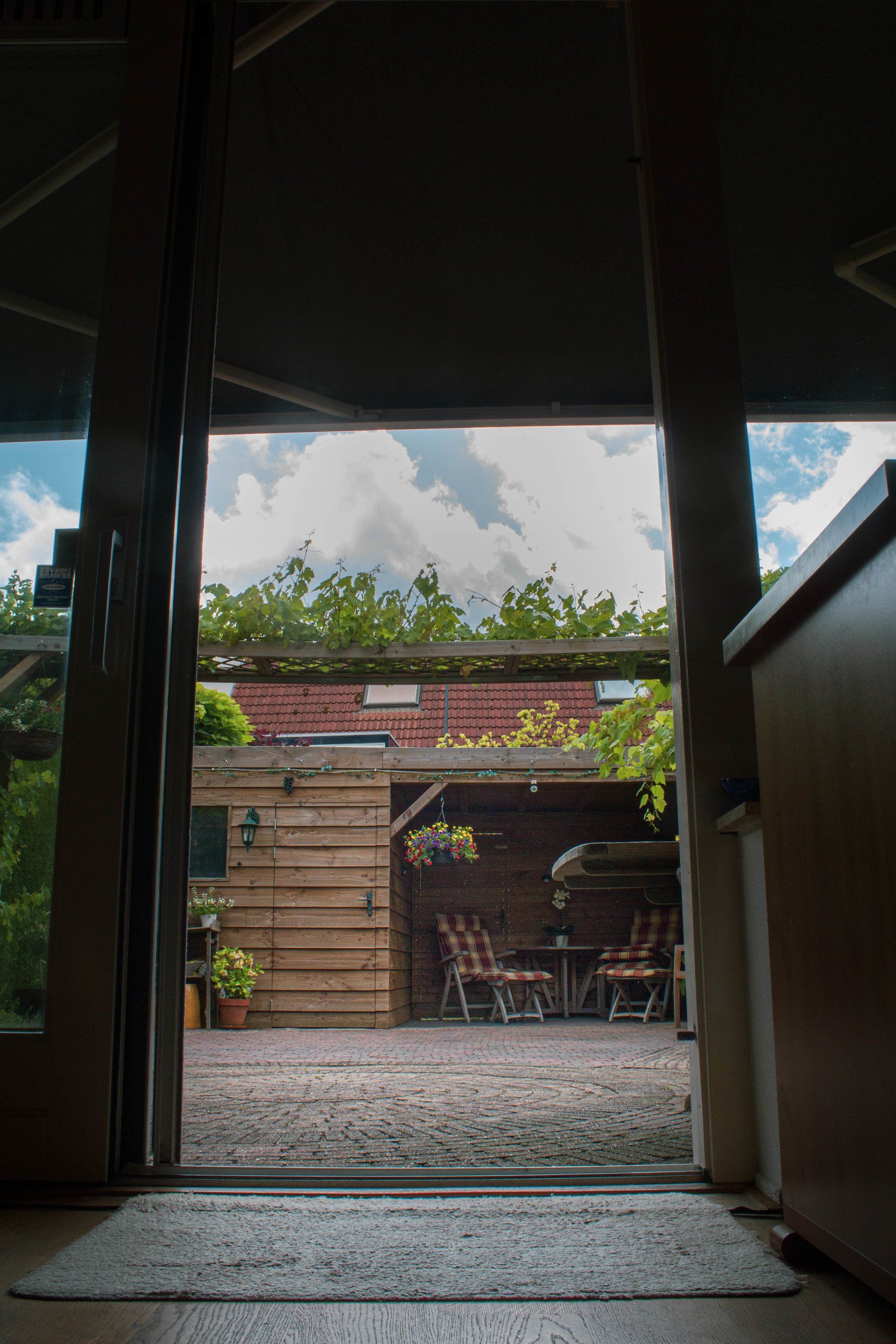 Uitzicht op de achtertuin vanuit binnen. Bewolkte lucht zichtbaar door de open deur. Buitenmeubilair en houten constructies zijn zichtbaar.
