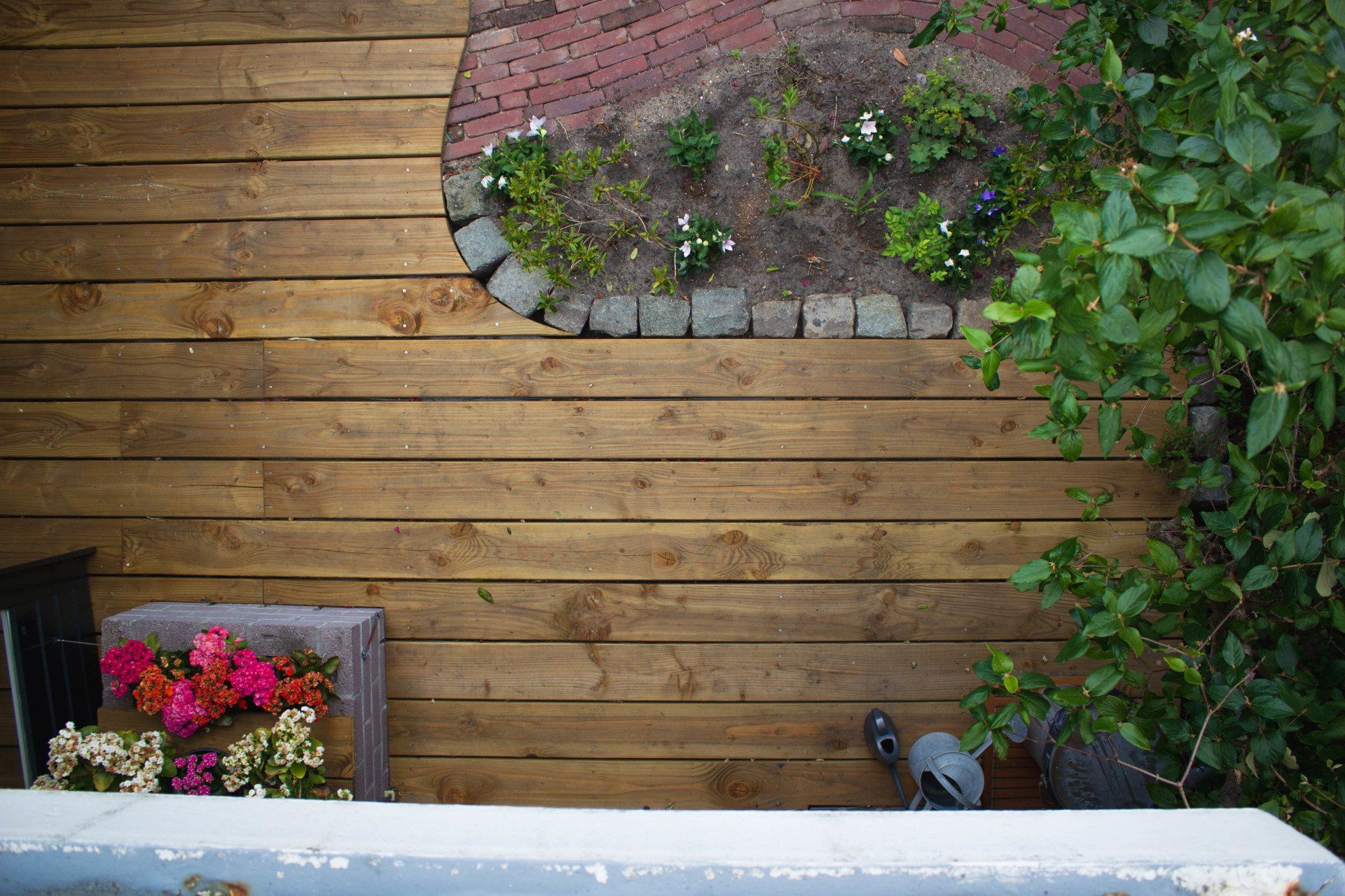 Houten terras met een bloembed en een bakstenen pad. Bloemen in een plantenbak en groen aan de rechterkant.