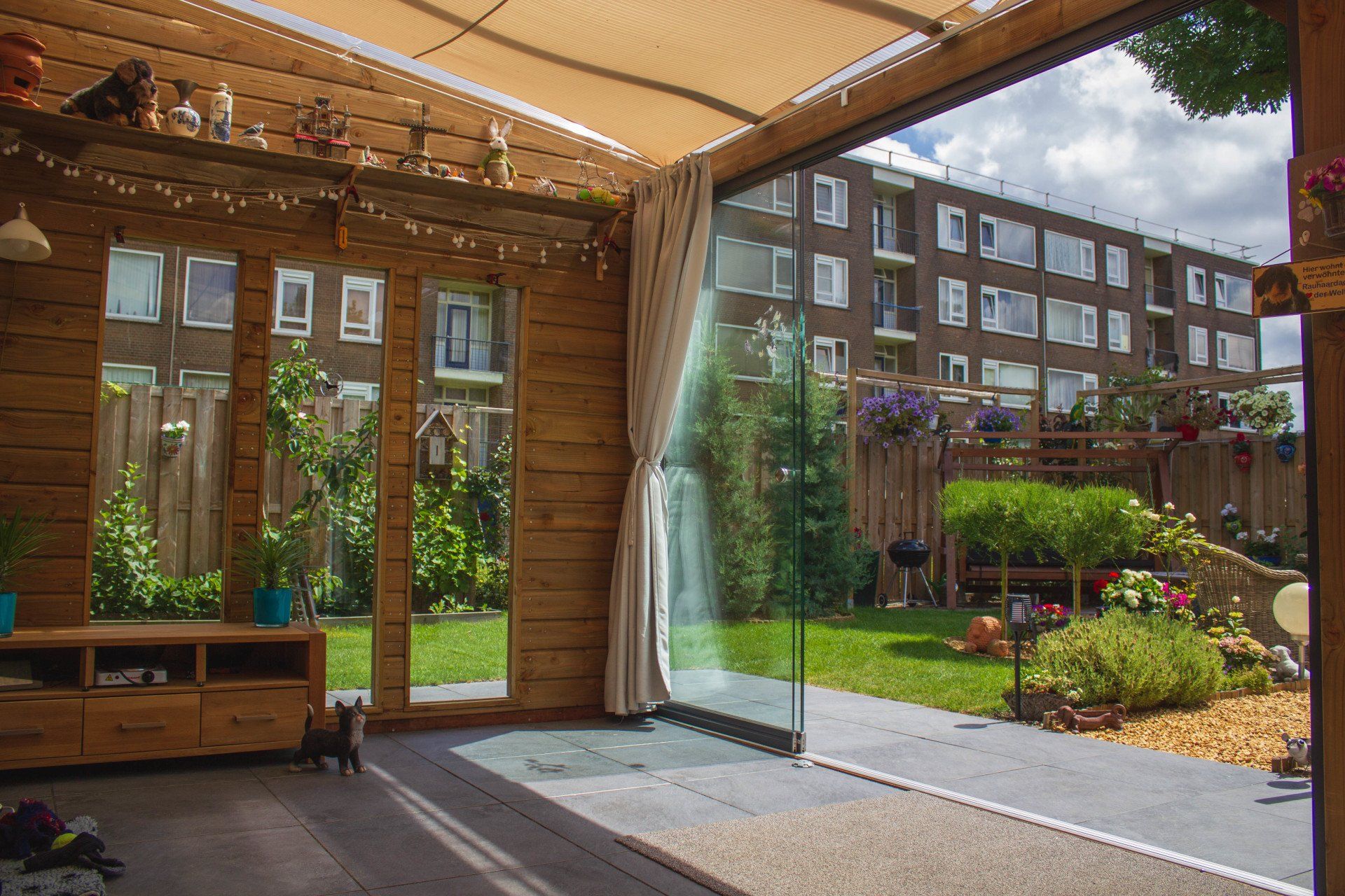 Binnen- en buitenruimte met een glazen wand en uitzicht op een weelderige achtertuin met een flatgebouw op de achtergrond.
