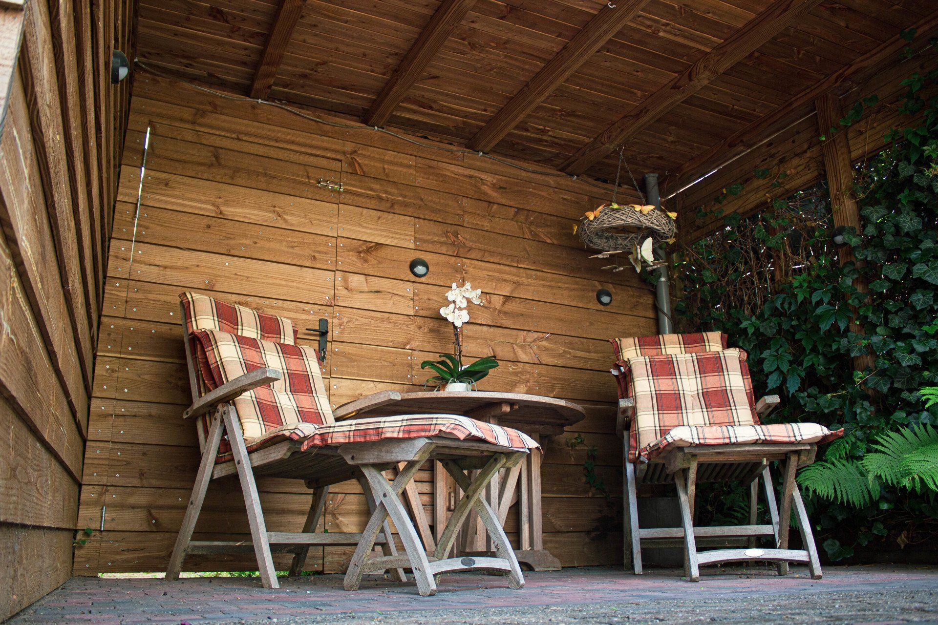 Twee houten stoelen met kussens met een patroon aan een klein tafeltje onder een overdekt terras met houten lambrisering.