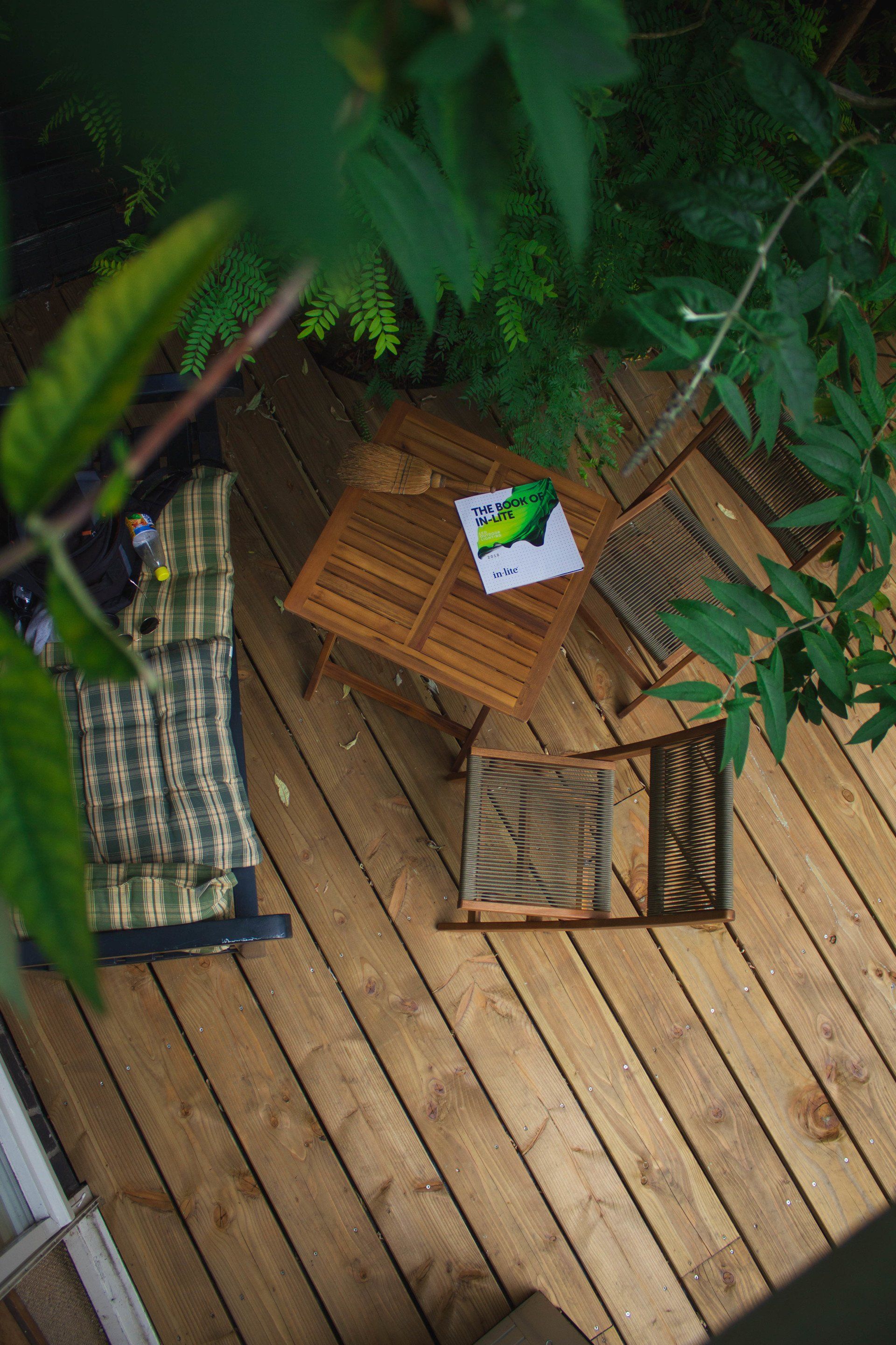 Bovenaanzicht van een houten terras met een klein tafeltje en twee stoelen, omgeven door groen, met een boek op het tafeltje.