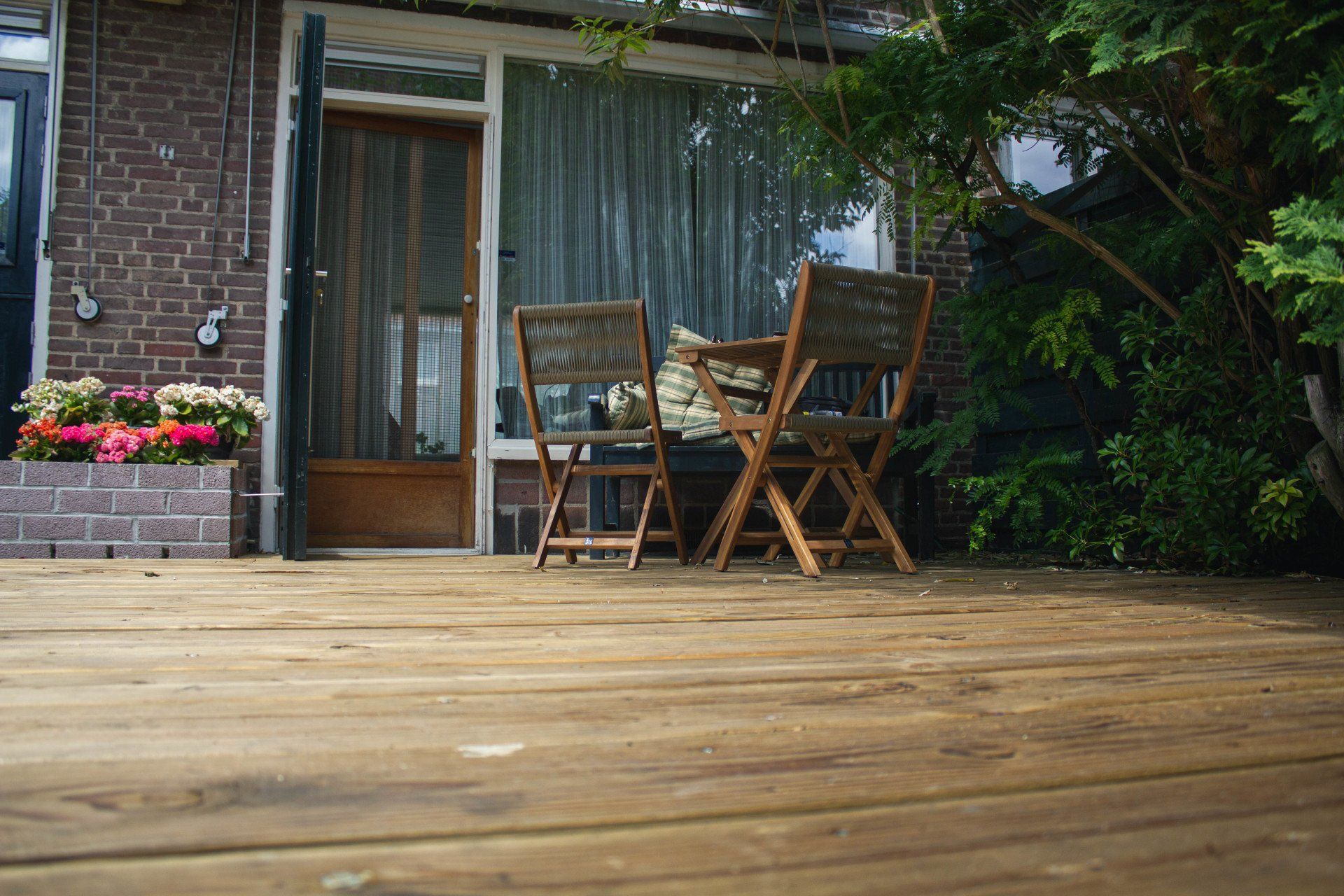 Houten terras met twee stoelen tegenover een deur, vlakbij een bakstenen gebouw met bloemen.