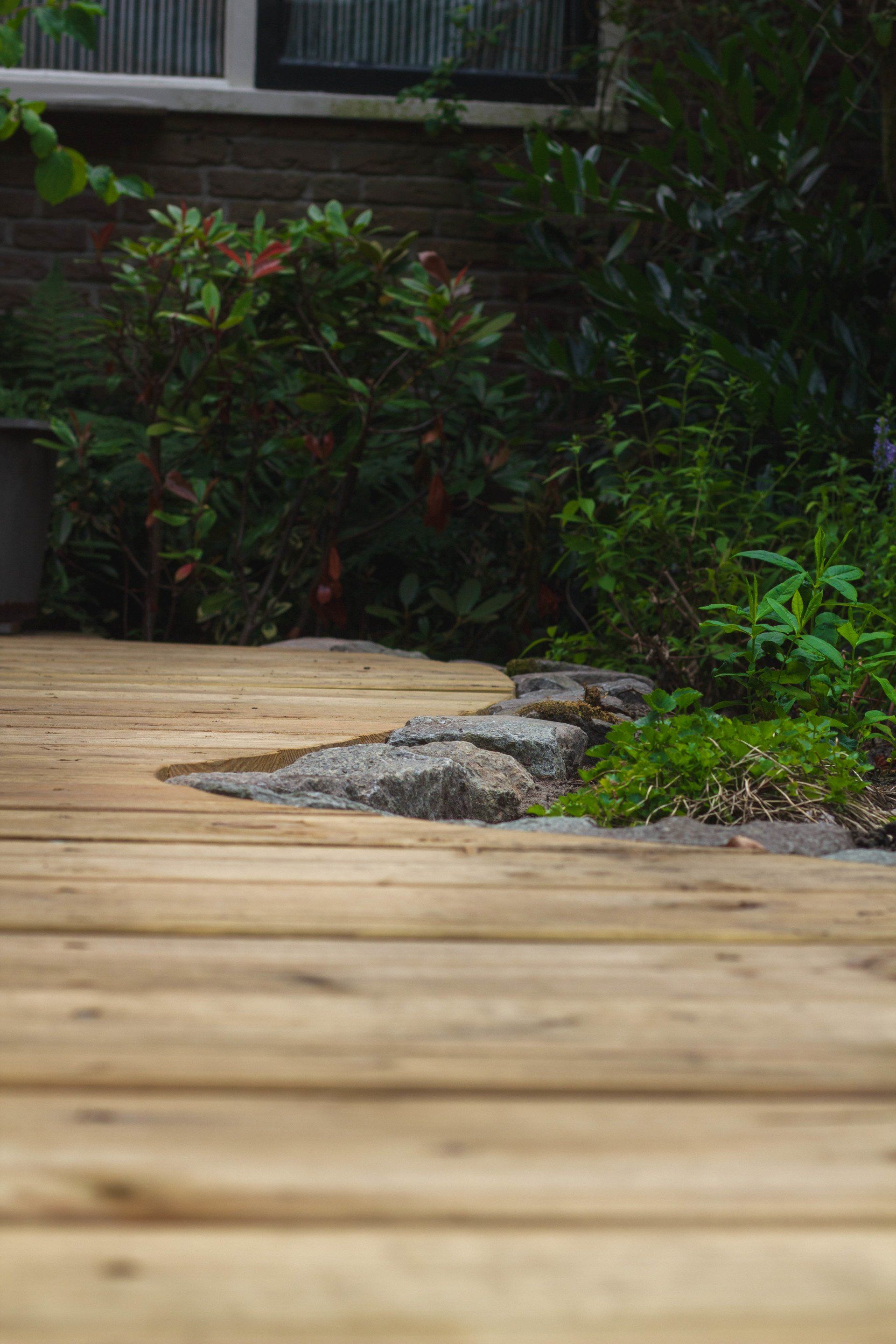Een houten terras leidt naar een bloembed met stapstenen, groen en een bakstenen muur.