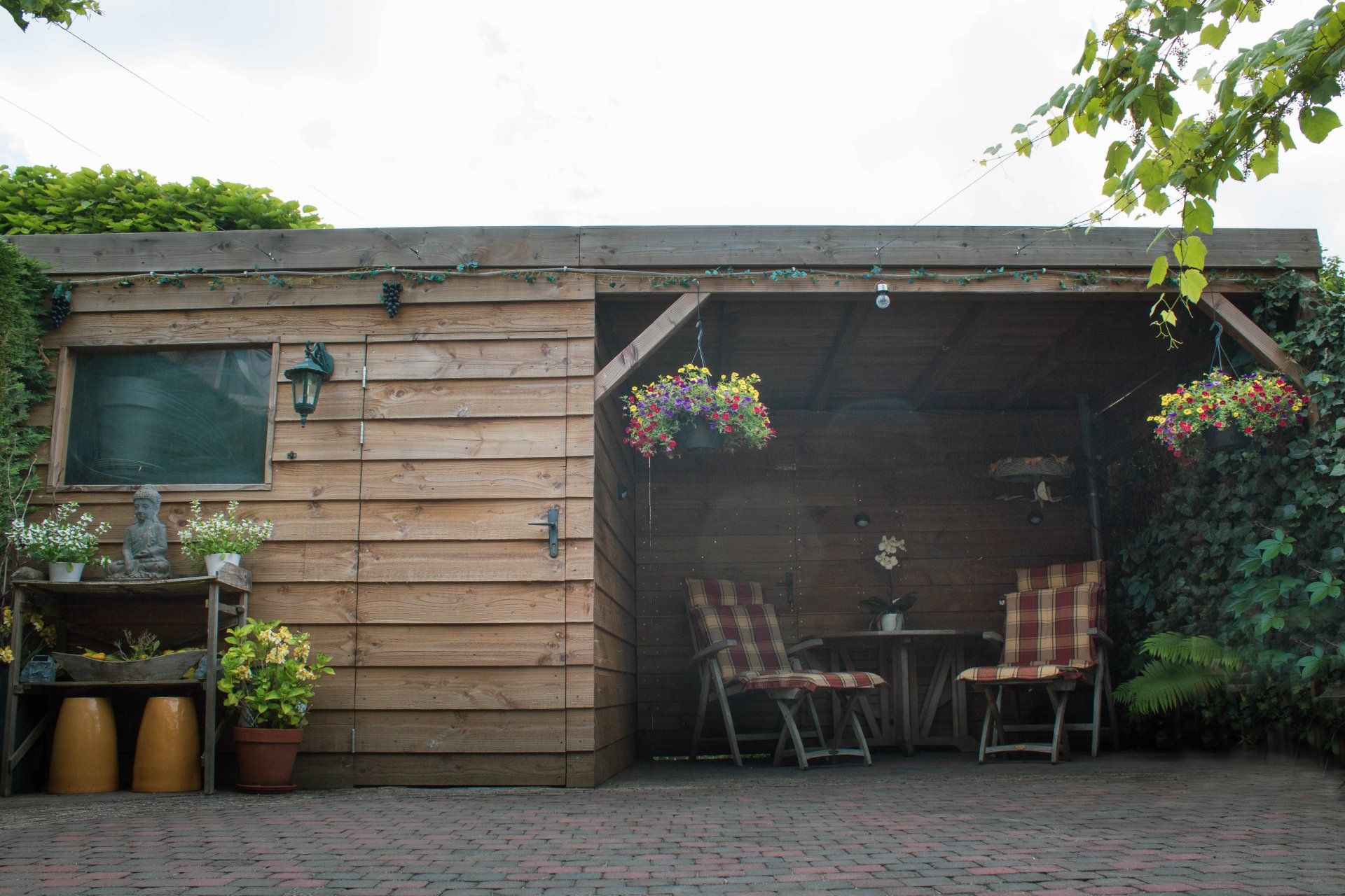 Houten schuur met terrasmeubilair; hangende bloemen, bakstenen vloer en planten.