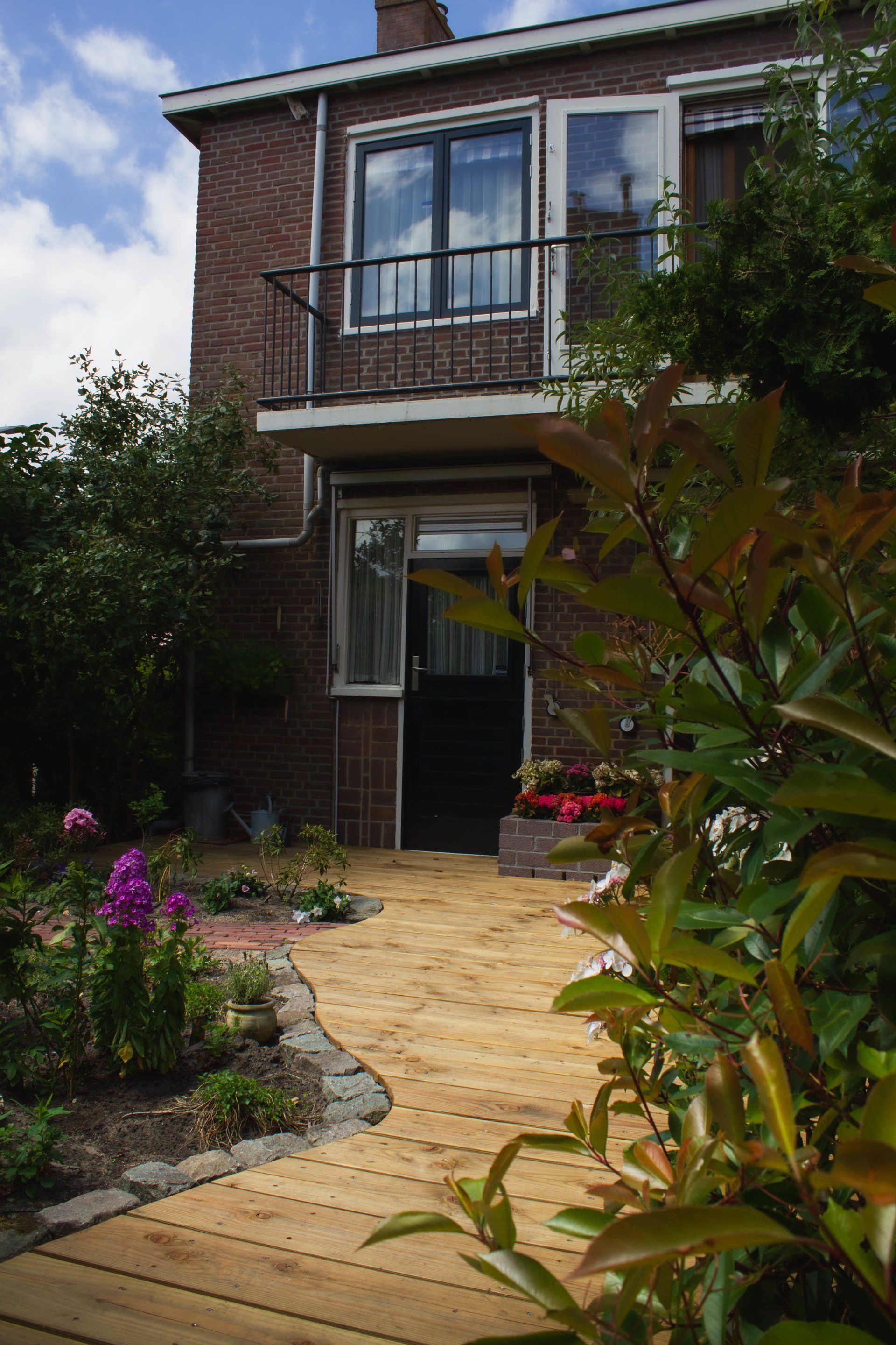 Een bakstenen huis met een klein balkon en een open deur, bereikbaar via een geel bakstenen pad door een tuin.