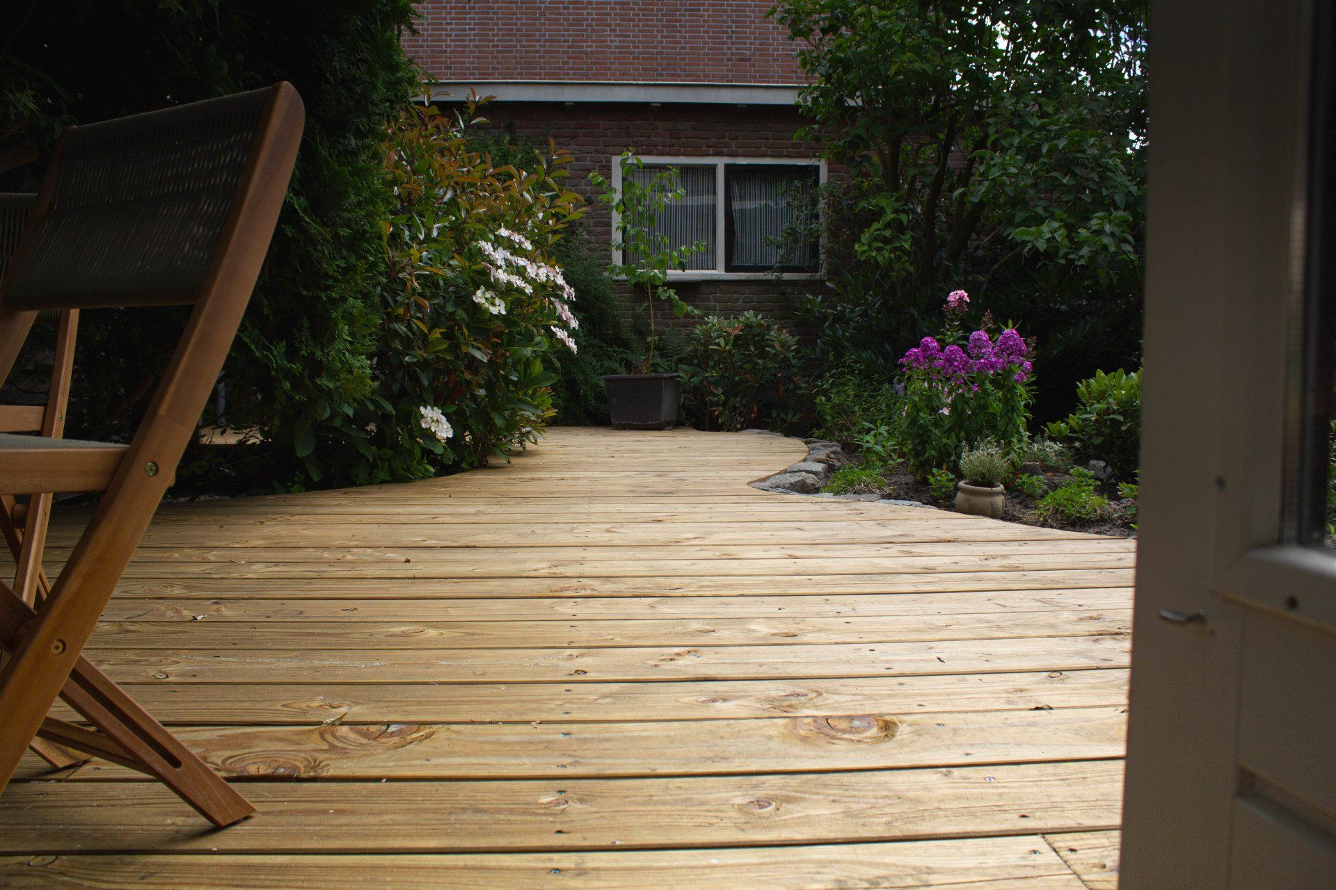 Een houten terras leidt naar een bakstenen gebouw met een raam, omgeven door groen en een stoel.