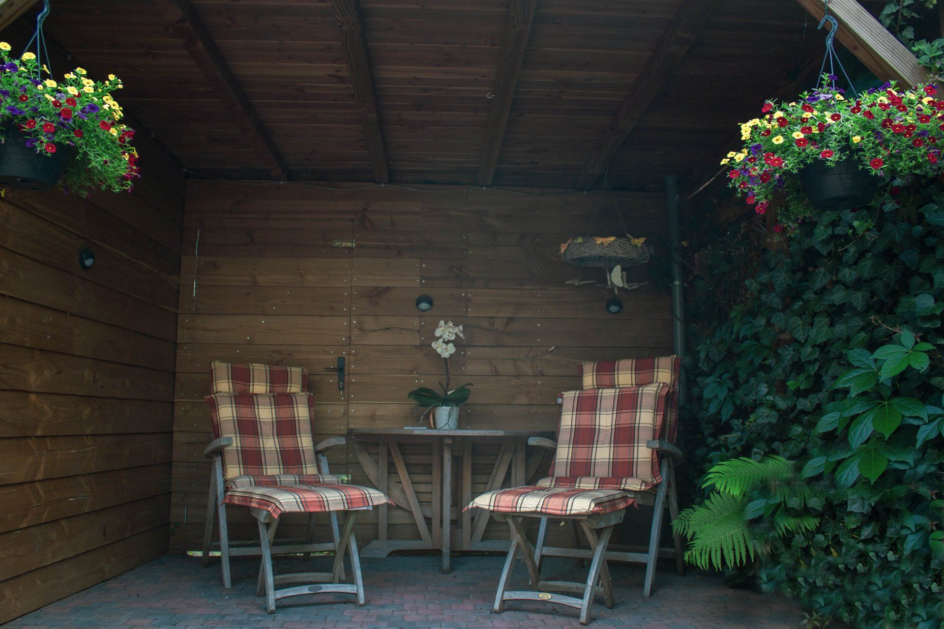 Twee stoelen met roodgeruite kussens staan ​​tegenover een kleine tafel in een houten afdakje, waaraan bloembakken hangen.