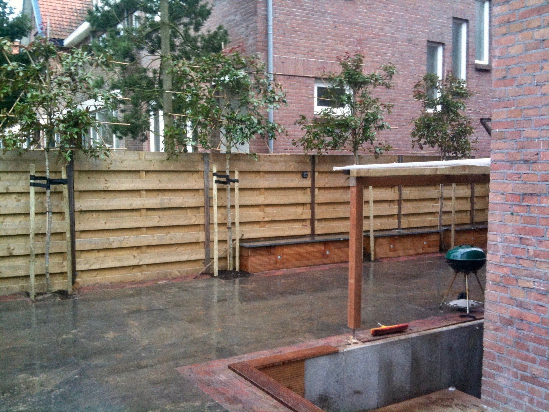 Achtertuin met een houten schutting, bomen, een kleine barbecue en een gedeeltelijk overdekt terras.
