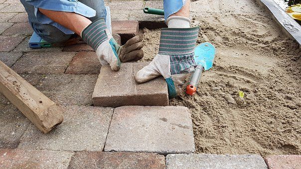 Een persoon met handschoenen aan legt stenen met zand in een buitenomgeving.
