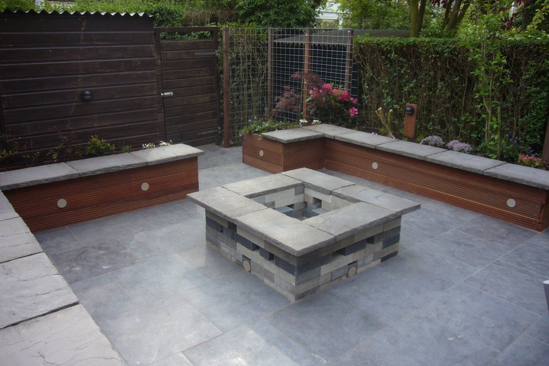 Terras met vuurkuil, ingebouwde houten banken, grijze natuurstenen bestrating en een donkere houten schutting als achtergrond.