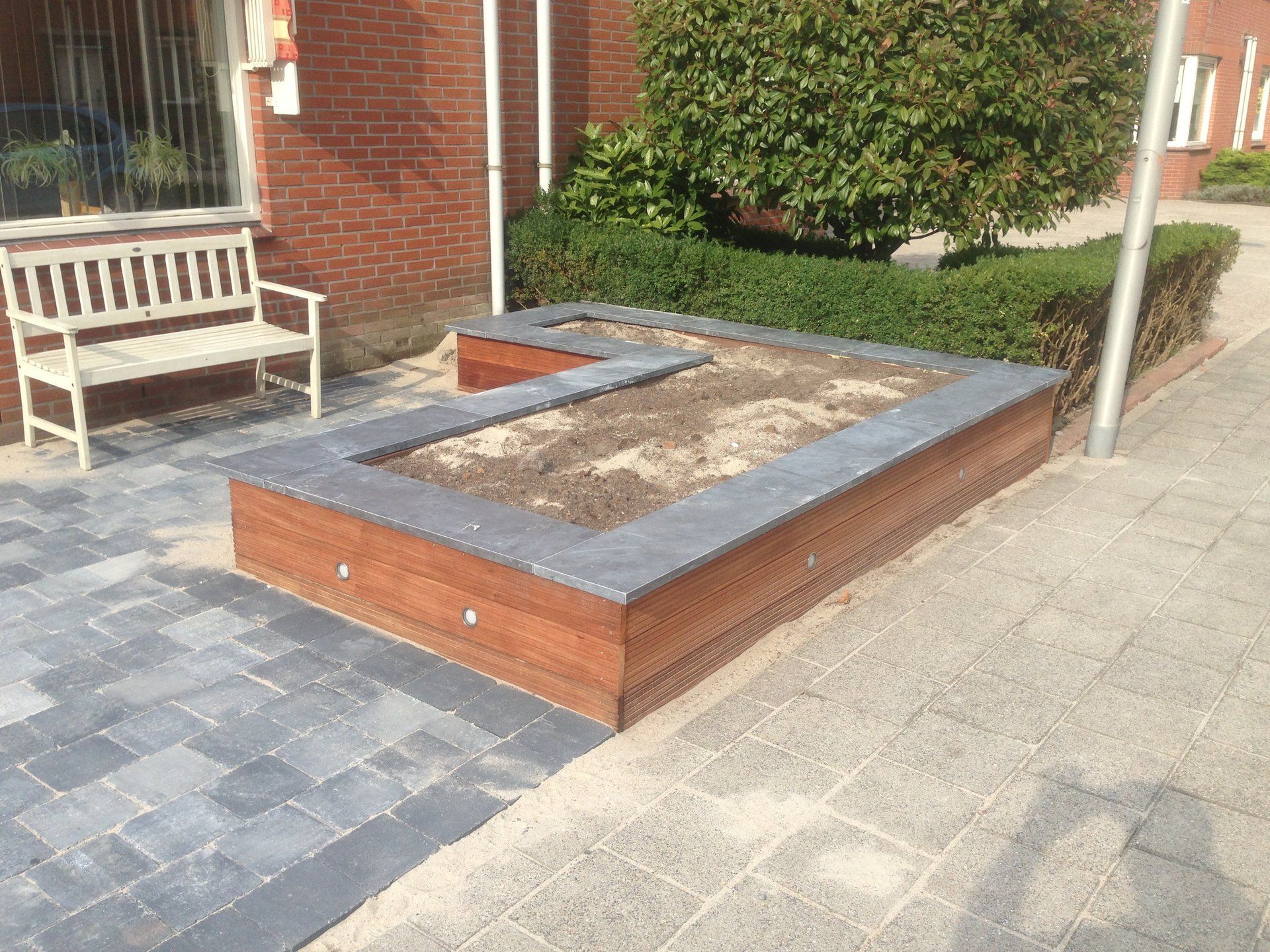 Verhoogd bloembed met een grijze rand en een bruine bakstenen basis op een bakstenen stoep naast een witte bank.