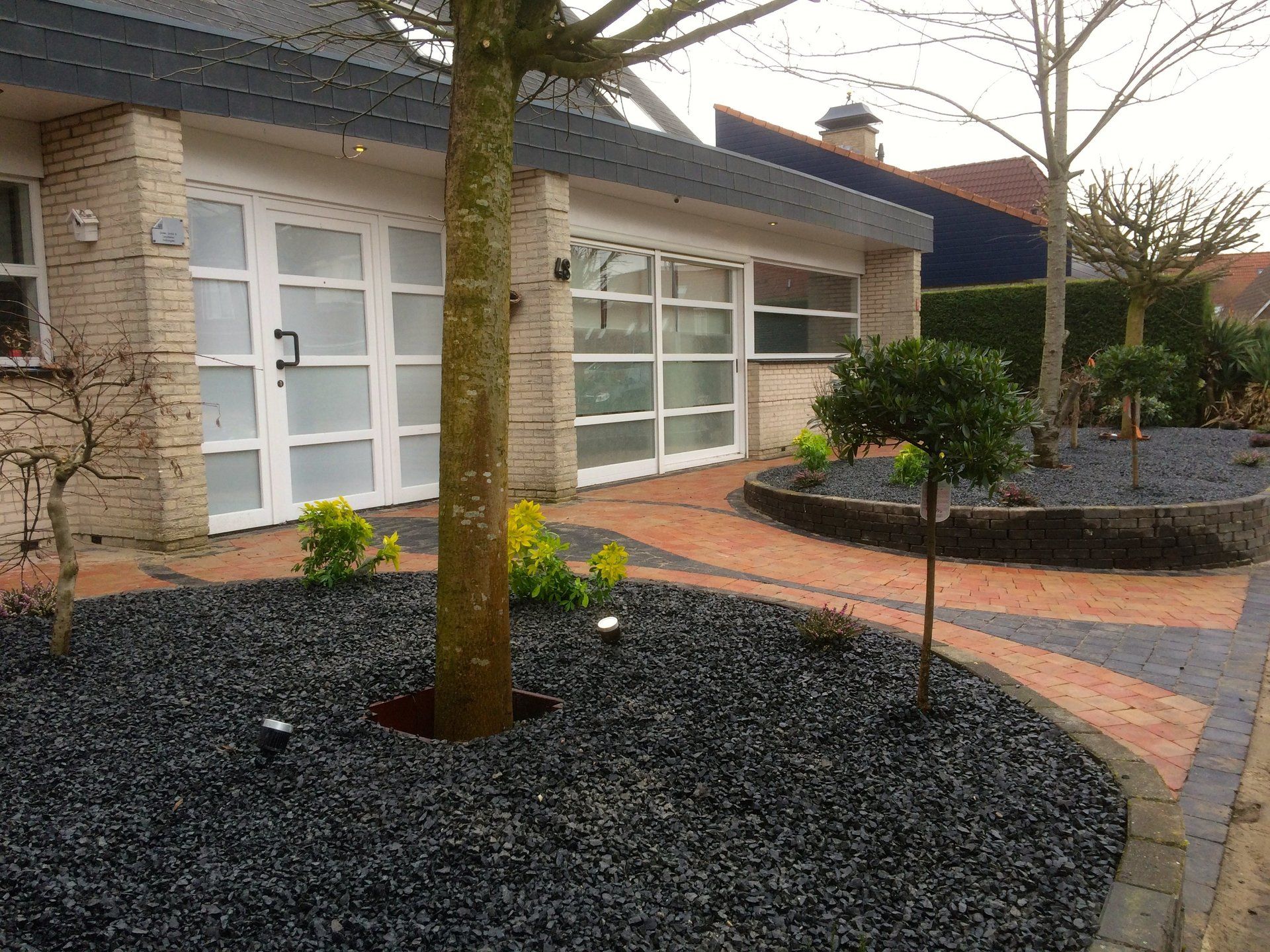 Een moderne woning met een zwartstenen perk in de voortuin, bomen en een bakstenen pad.