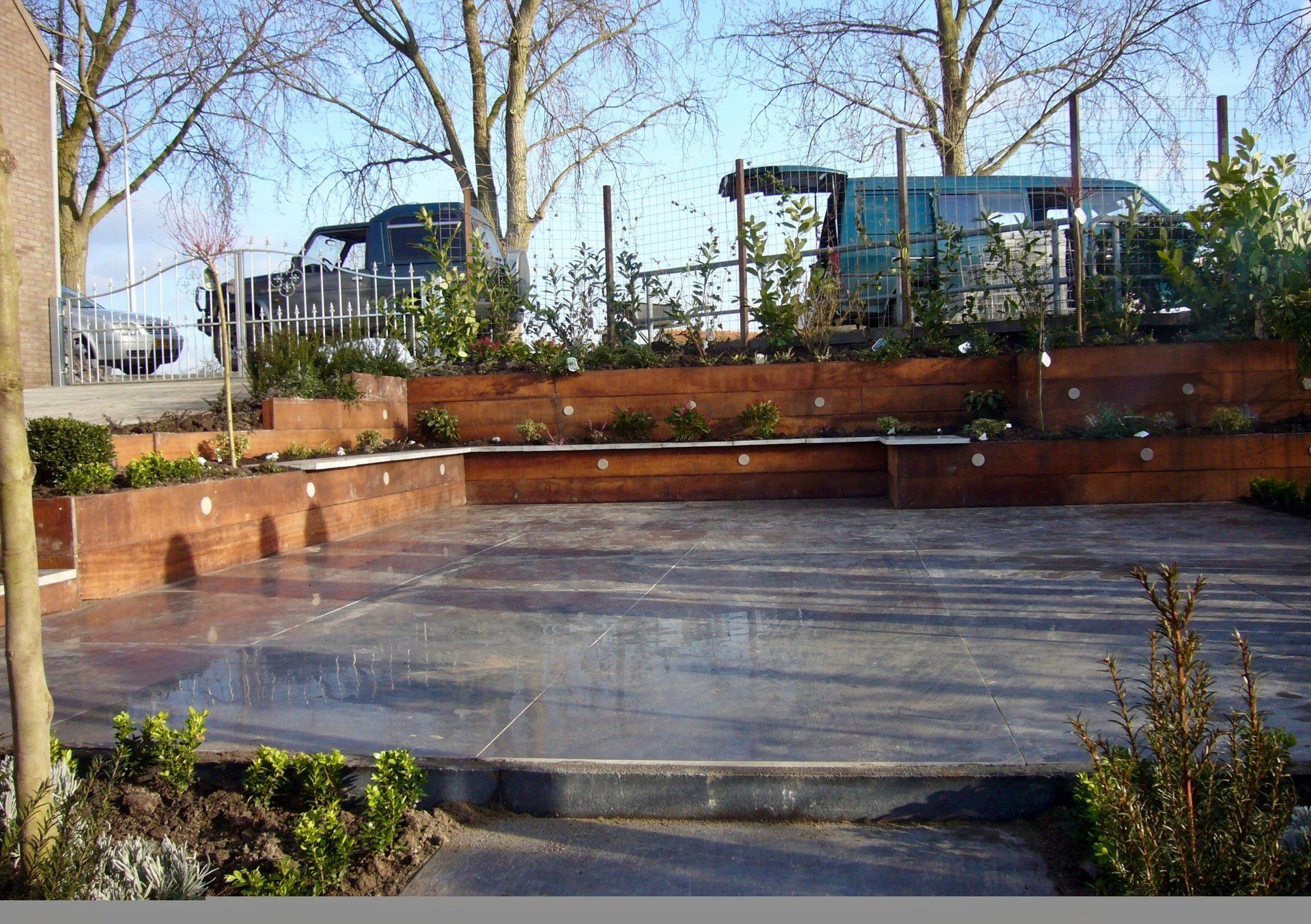 Verhoogde houten plantenbakken met groen omringen een betonnen terras, met voertuigen en bomen op de achtergrond.
