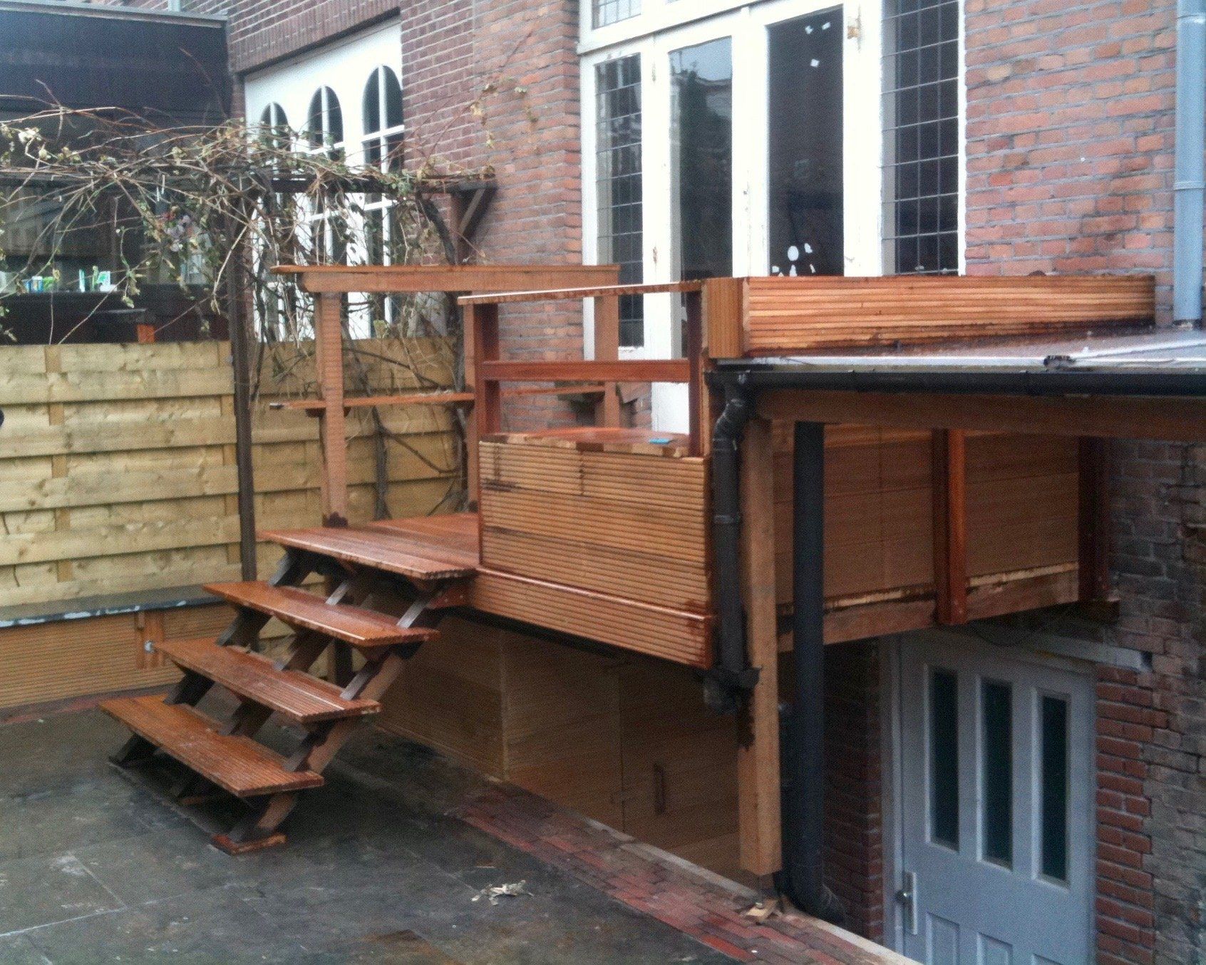 Houten terras met trappen, bevestigd aan een bakstenen gebouw; onder het terras is een deur zichtbaar.