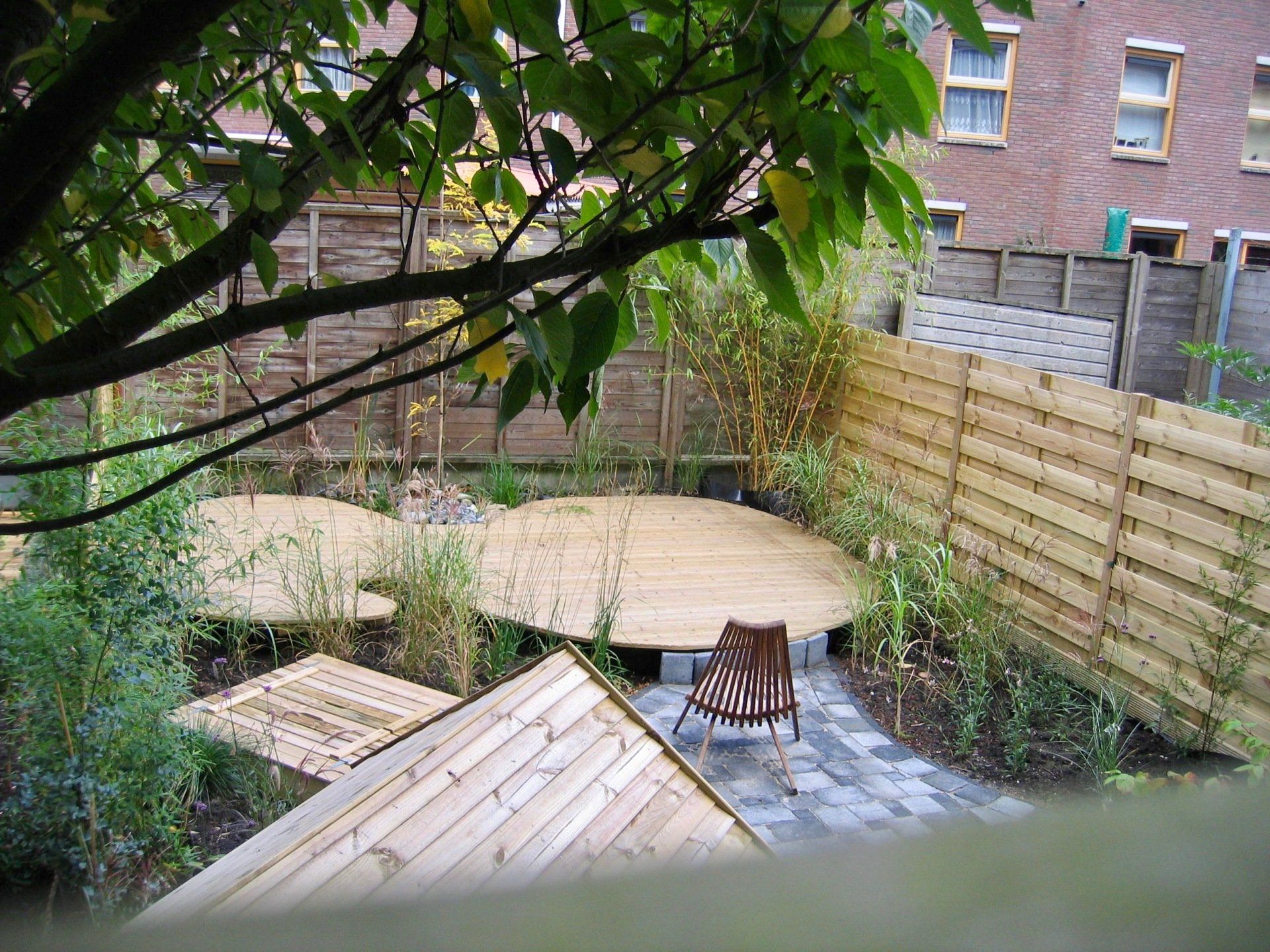 Achtertuin met bamboe schutting, houten platforms en een stoel op een stenen pad, met een gebouw op de achtergrond.