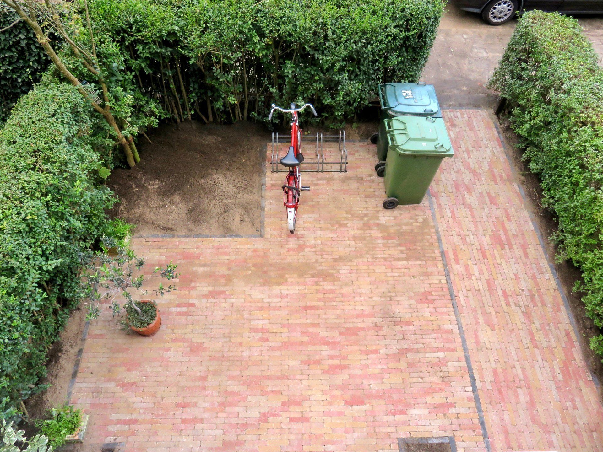 Terras van rode bakstenen met groene afvalbakken, fiets en hagen.