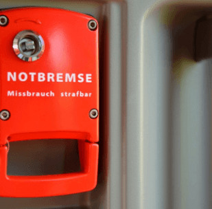 Roter Notbremshebel an einer Metallwand, deutscher Text: 