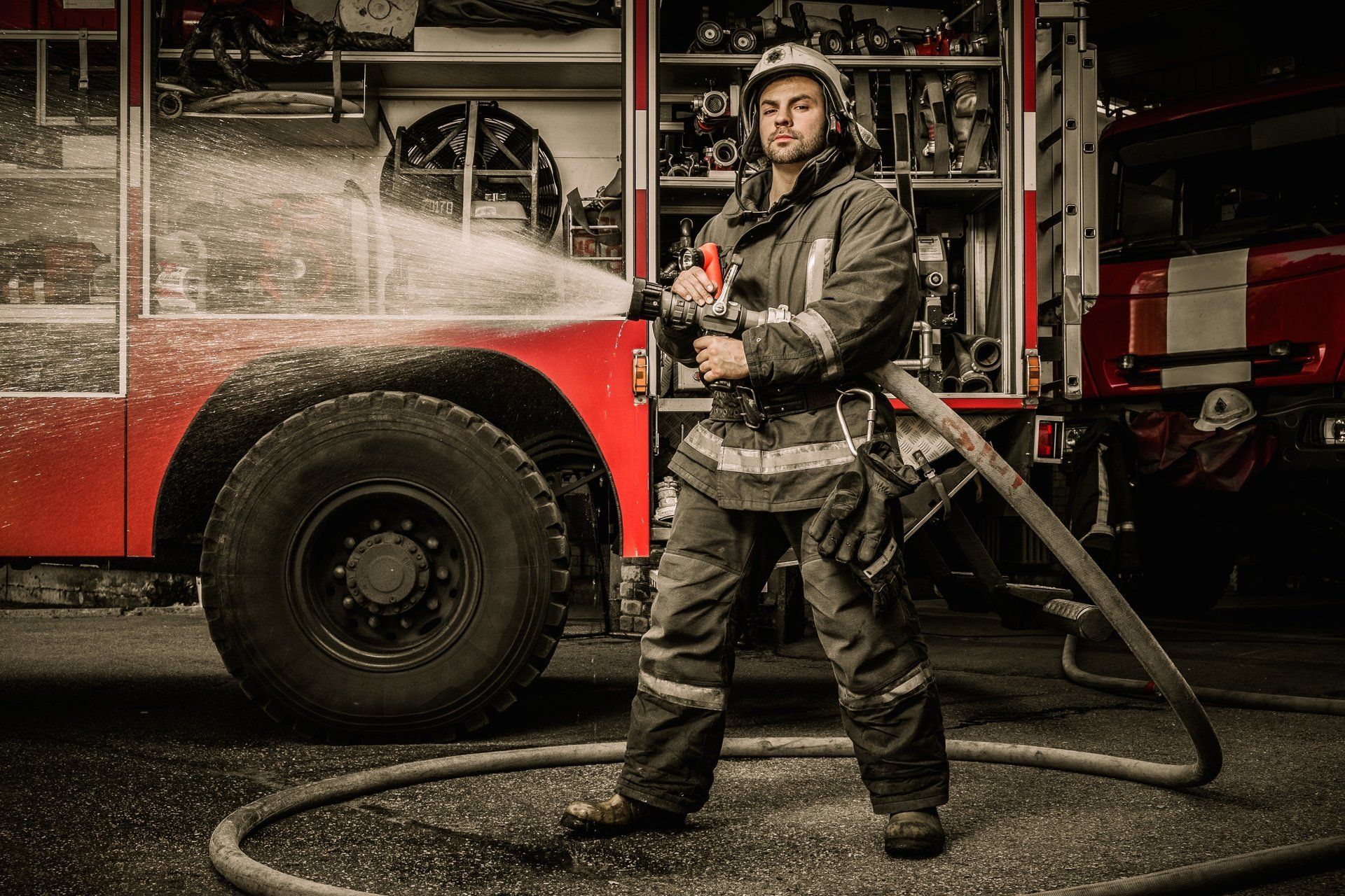 Ein Feuerwehrmann spritzt Wasser aus einem Schlauch; im Hintergrund ist ein rotes Feuerwehrauto zu sehen.