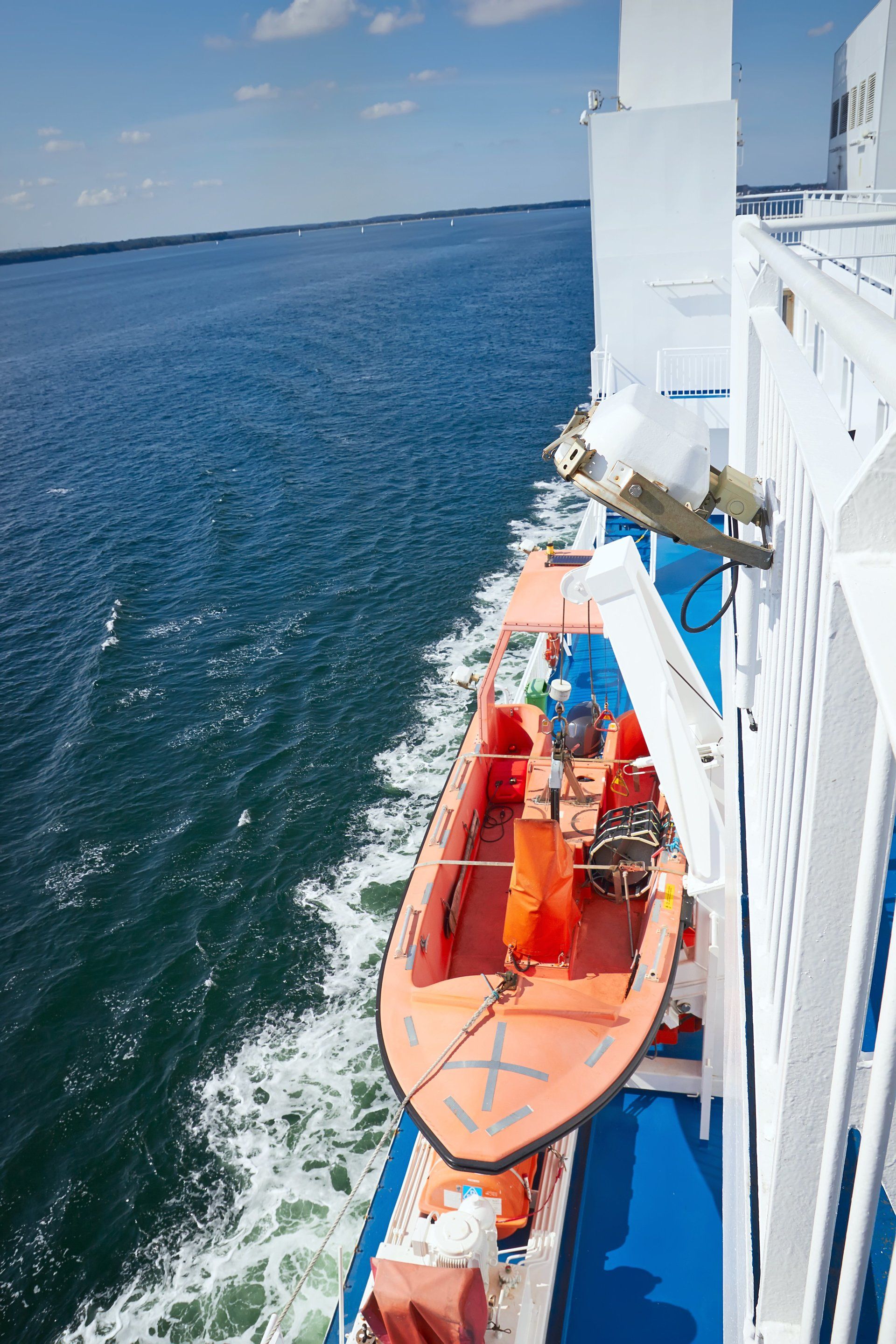 Ein orangefarbenes Rettungsboot hängt an der Seite einer weißen Fähre an einem sonnigen Tag mit blauem Wasser und Himmel.