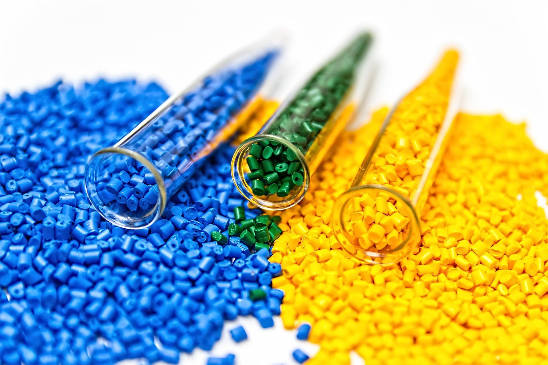Blaue, grüne und gelbe Kunststoffpellets in Reagenzgläsern und verstreut auf einer weißen Oberfläche.