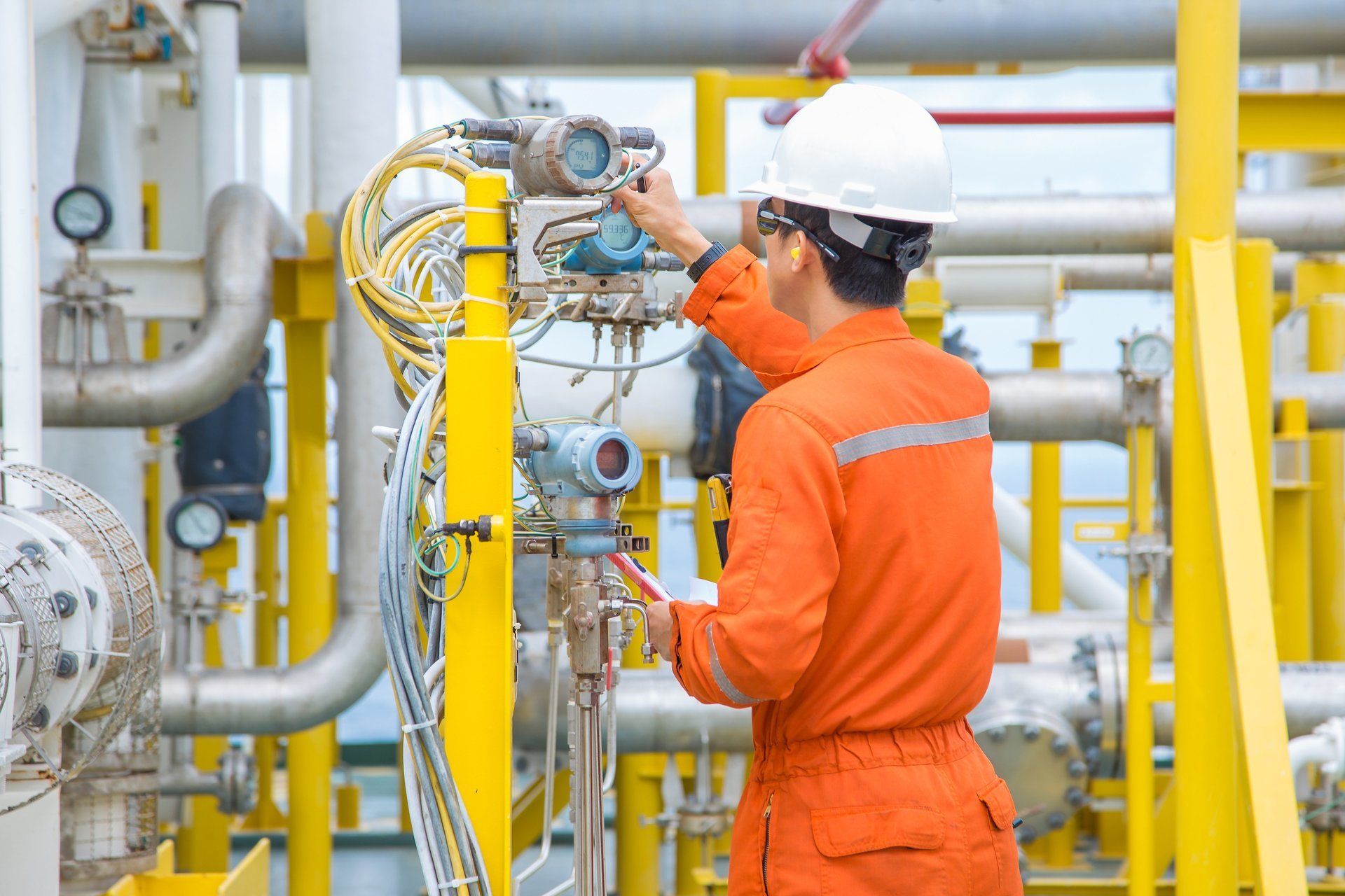 Ein Arbeiter in orangefarbenem Overall und Schutzhelm inspiziert eine Anlage auf einer Industrieplattform.