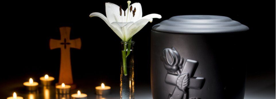 Candele e una croce di legno accanto a un fiore bianco e un'urna nera su un altare commemorativo scuro