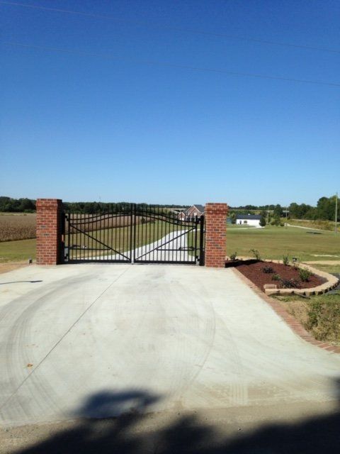Black iron gate set between brick columns, spanning a concrete driveway leading into a rural property.