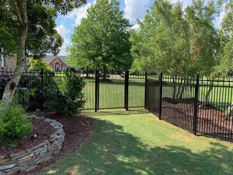 Black metal fence with gate in a yard, trees in the background, a stone wall on the left.