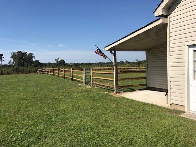 Lawn with a wooden fence, American flag, and a house with light tan siding under a blue sky.