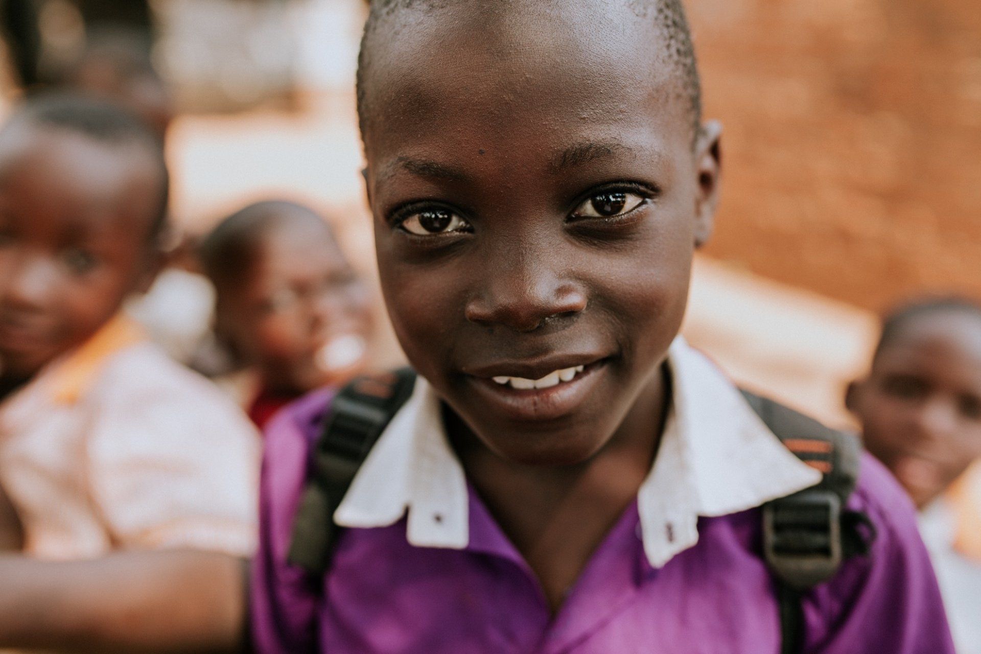 A young boy in a purple shirt with a backpack is smiling for the camera.
