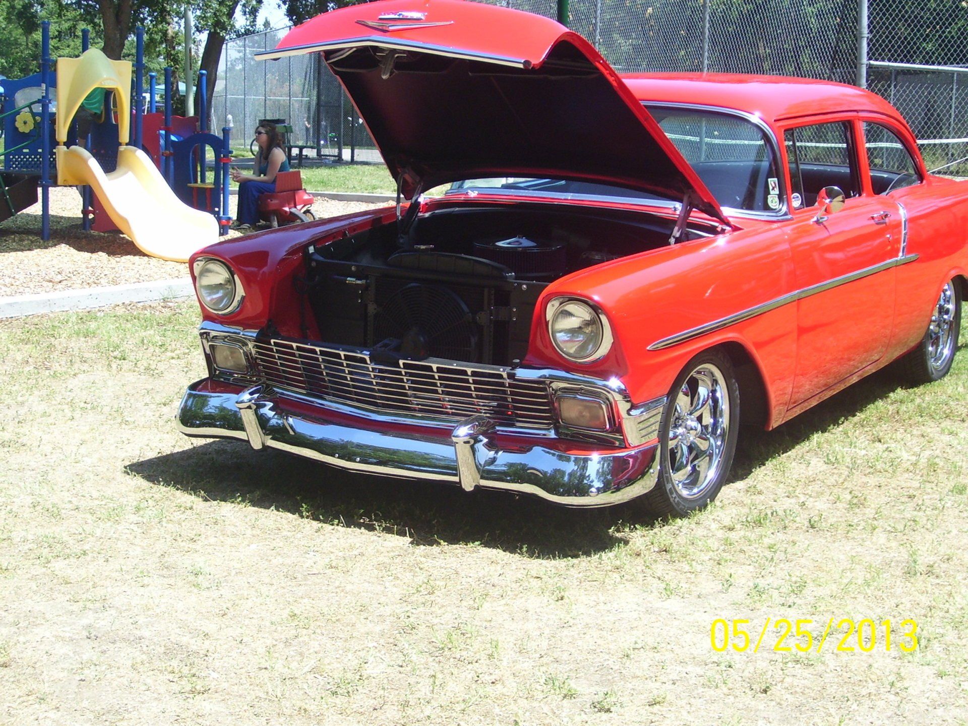 A red car with the hood up is parked in front of a playground