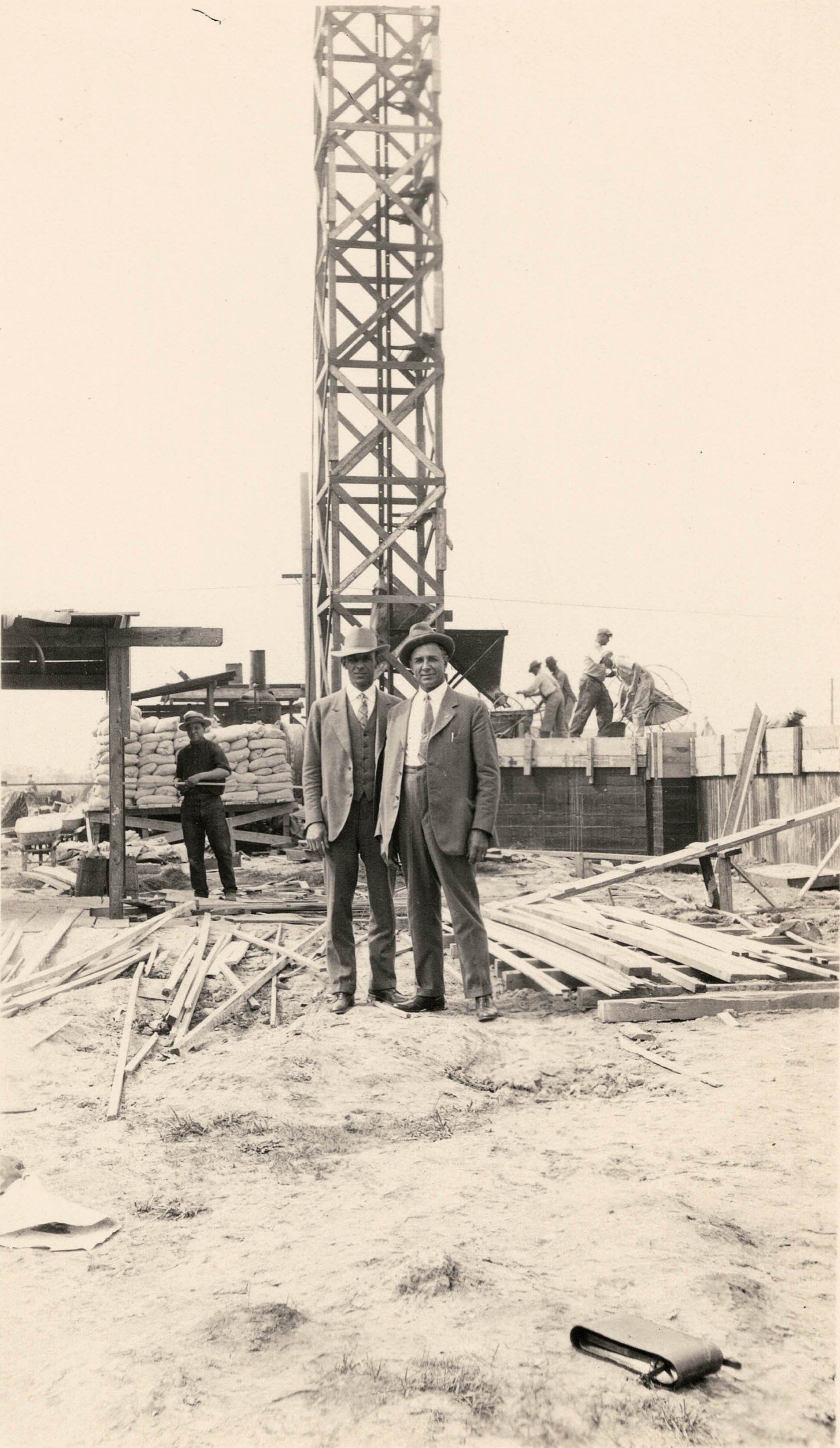 Two men are standing in front of a crane in a black and white photo.