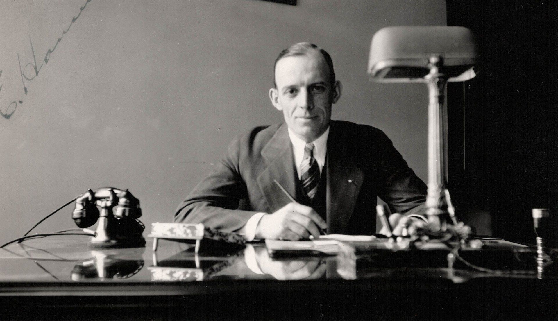 A man in a suit and tie is sitting at a desk with a lamp and a telephone