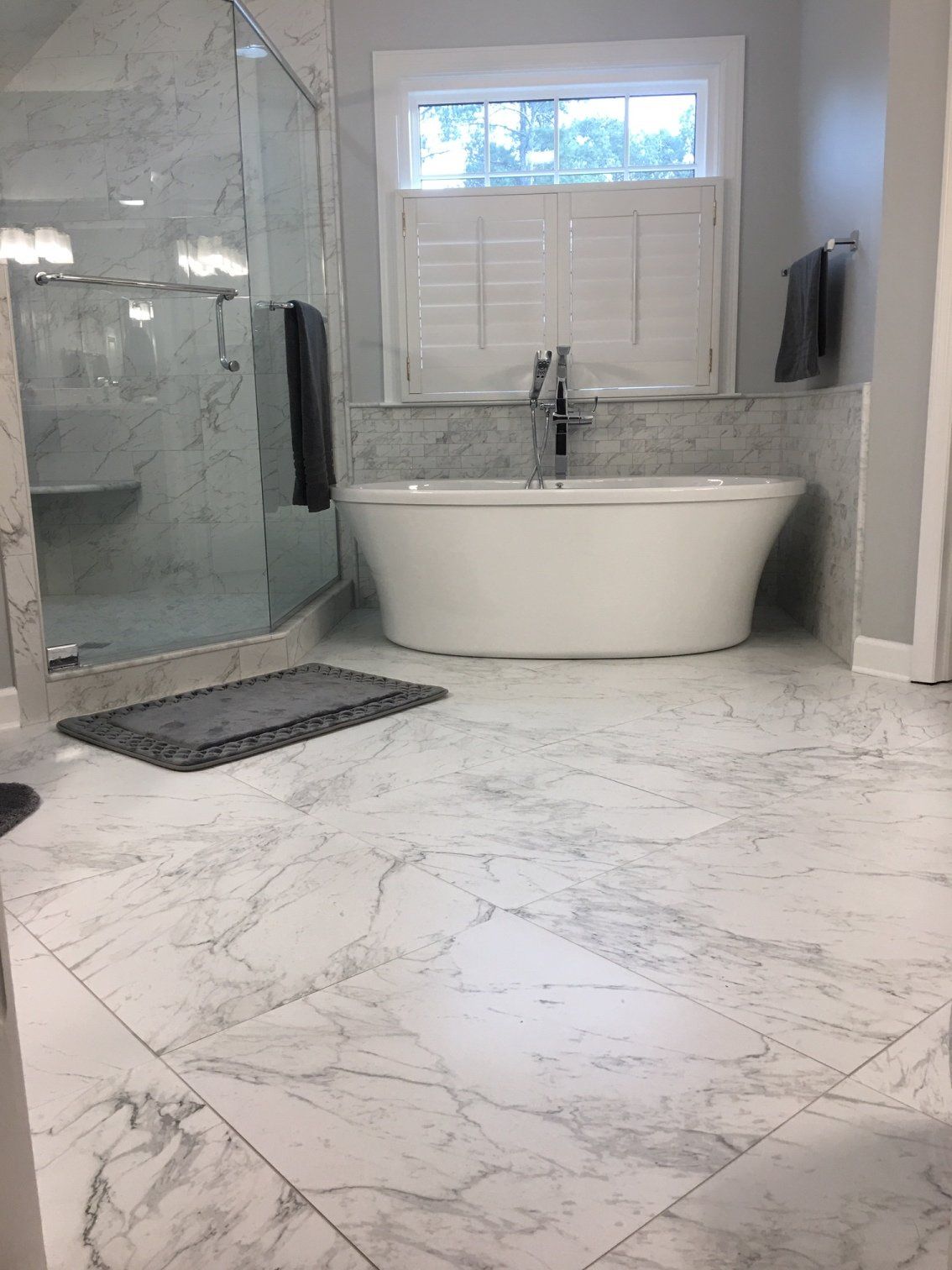 White marble-tiled bathroom with a freestanding tub, glass shower, and window with white shutters.