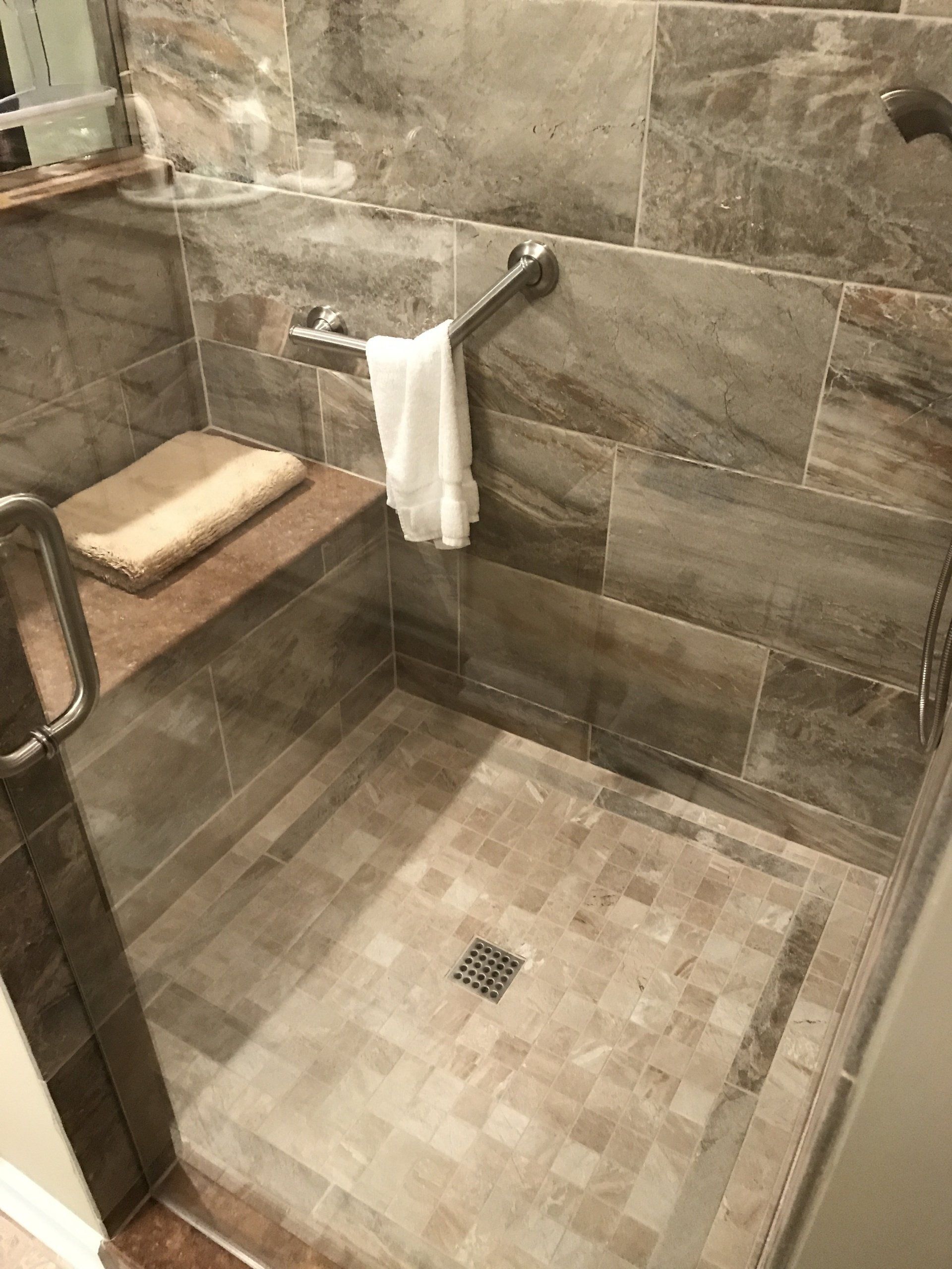 Accessible shower with grab bar, bench, and glass door. Beige tile, white towel.