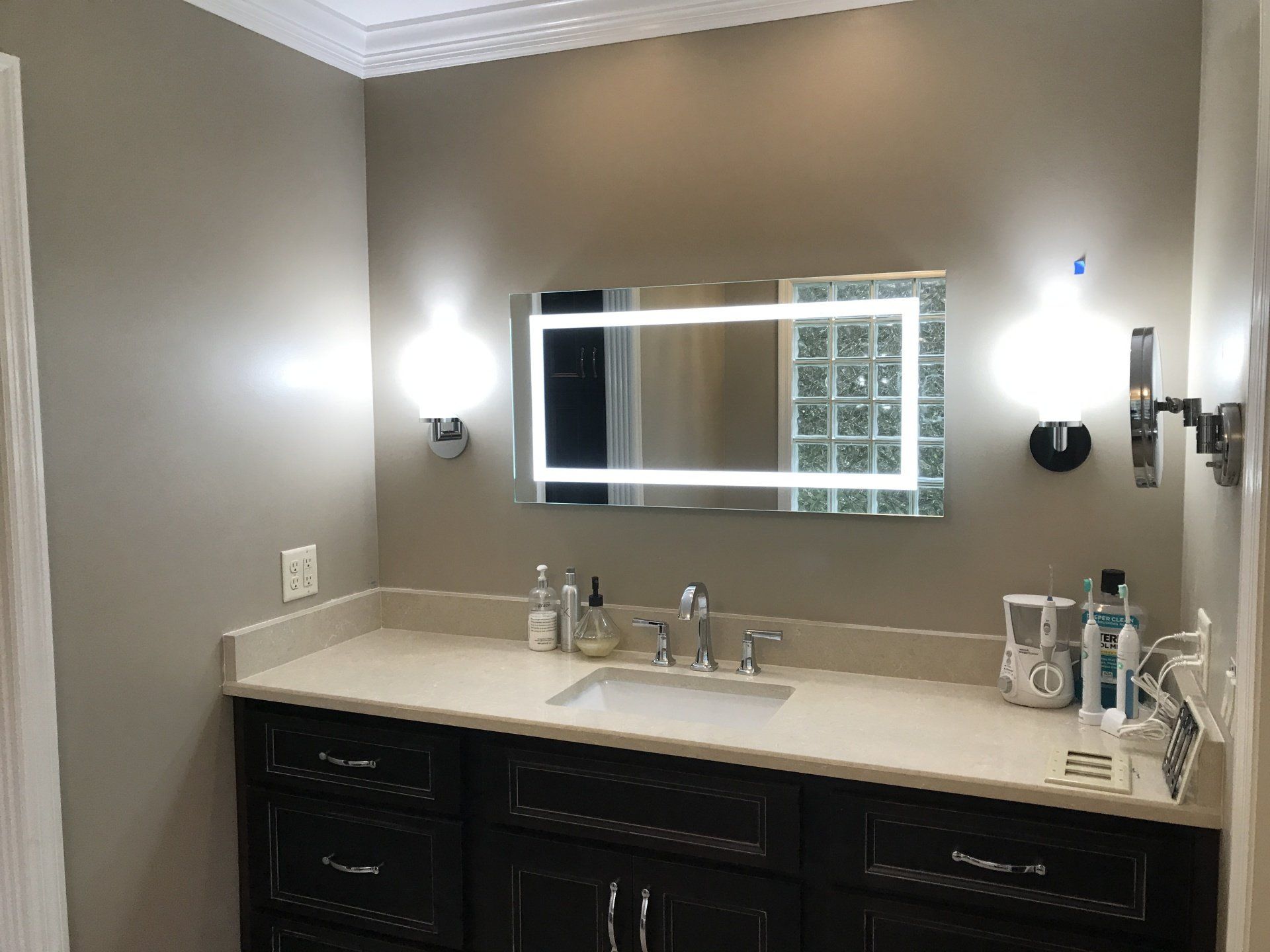 Bathroom vanity with a lighted mirror, sconces, dark cabinets, and a beige countertop.