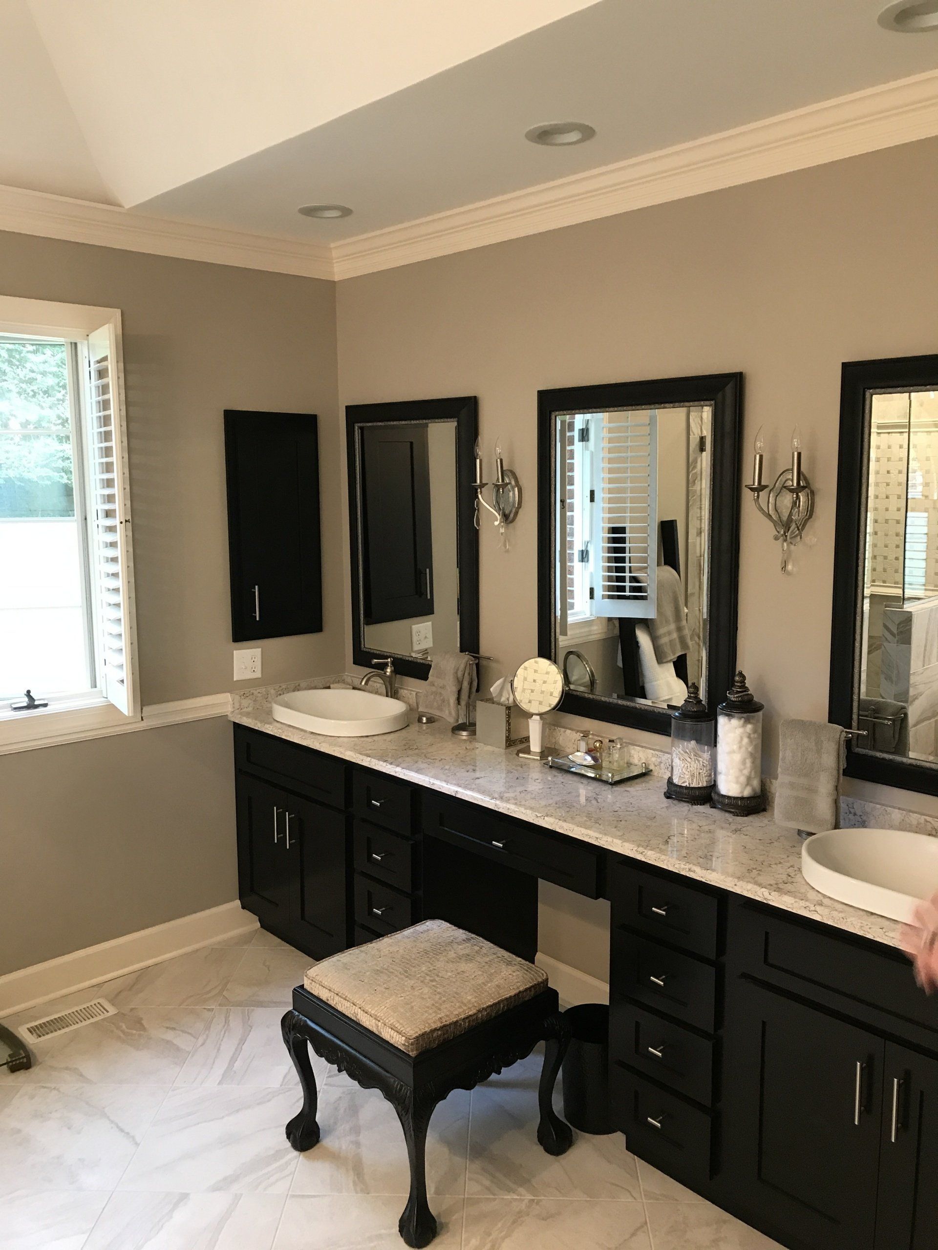 Bathroom with black vanities, marble countertops, and three mirrors; a white window, and a patterned stool.